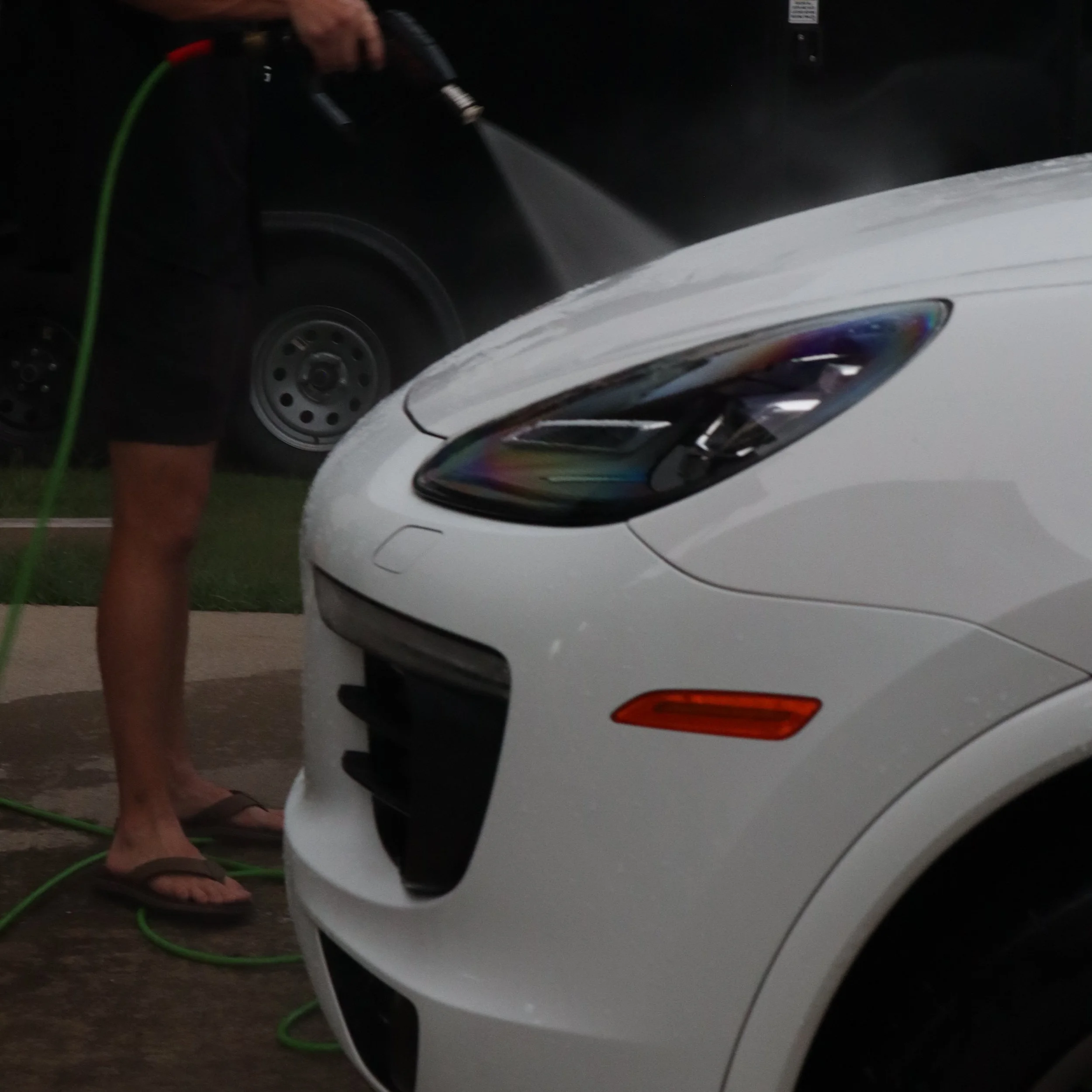 Person cleaning the front of a white car with a spray bottle, standing outdoors at night.