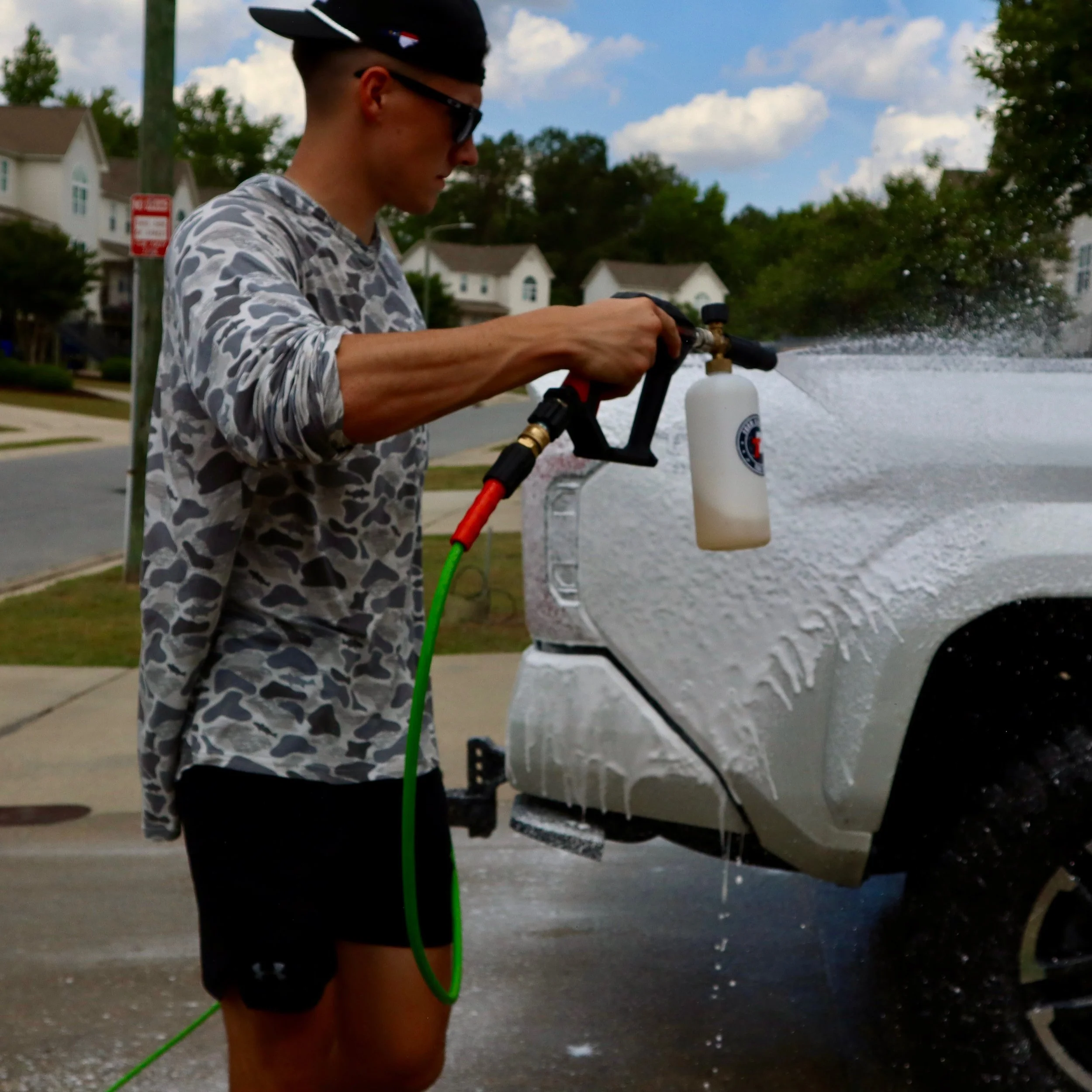 A person dressed in a camouflage long-sleeve shirt and shorts, wearing sunglasses and a helmet, is holding a pressure spray bottle and spraying water on a window or glass surface outdoor during the daytime.
