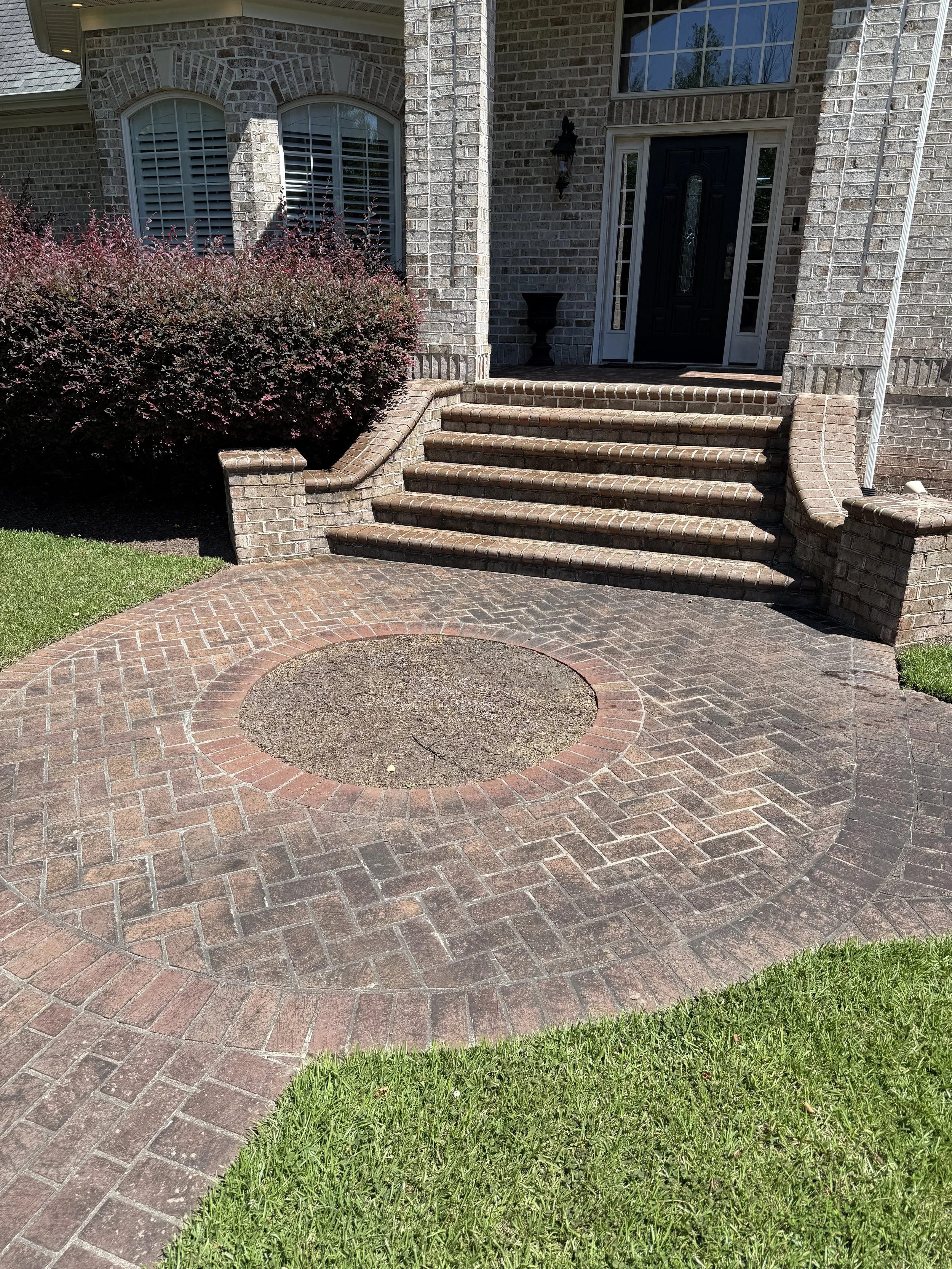 Brick front steps leading to a black front door of a house with large windows and a bush on one side, a circular brick patio in front, and green grass surrounding the area.