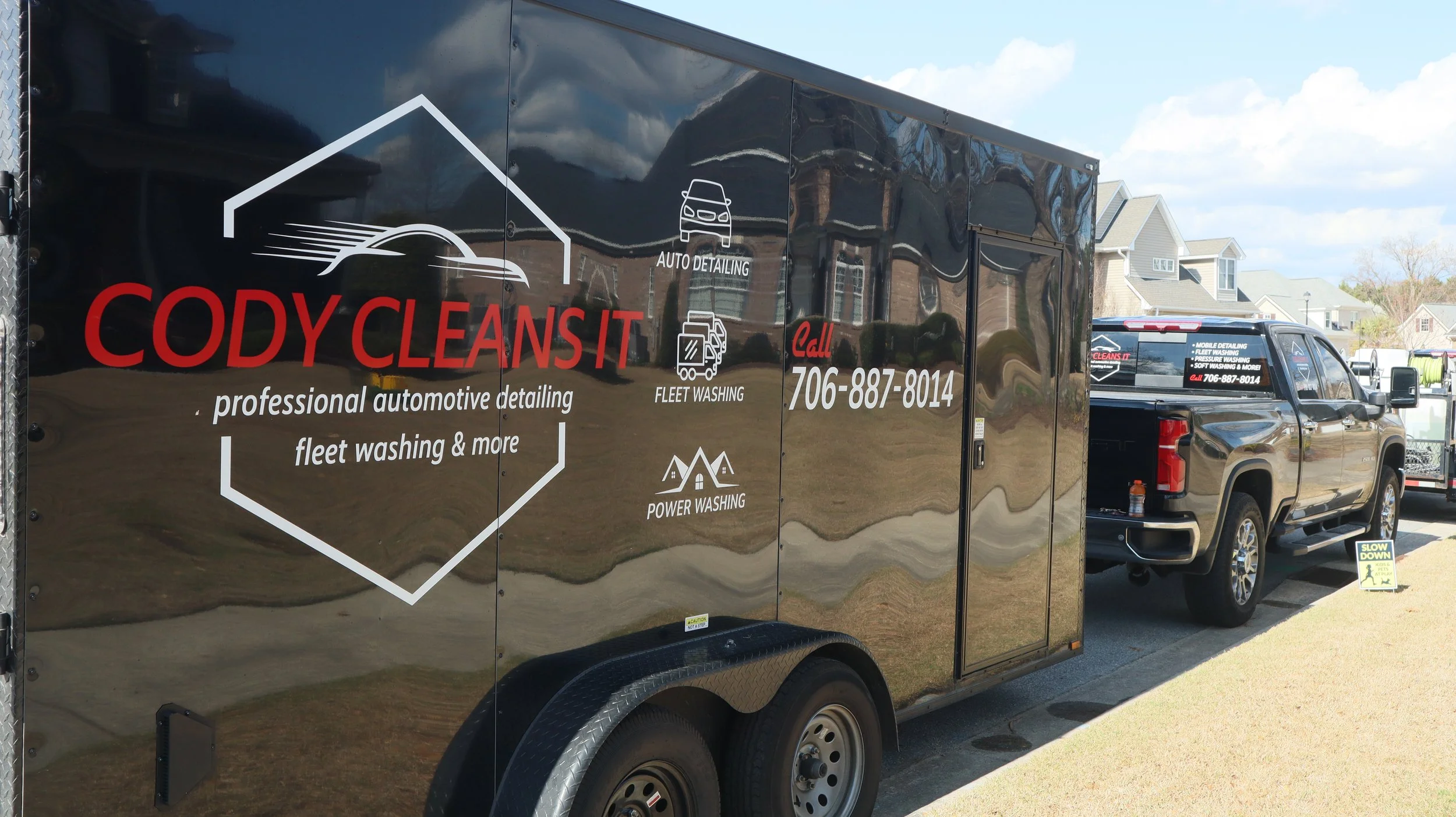 A black enclosed trailer with the logo and contact information for Cody Cleans It, a professional automotive detailing service, parked on a residential street. The trailer lists services such as auto detailing, fleet washing, and power washing. A black pickup truck is attached to the trailer, and a small yellow sign on the ground says "Slow Down."