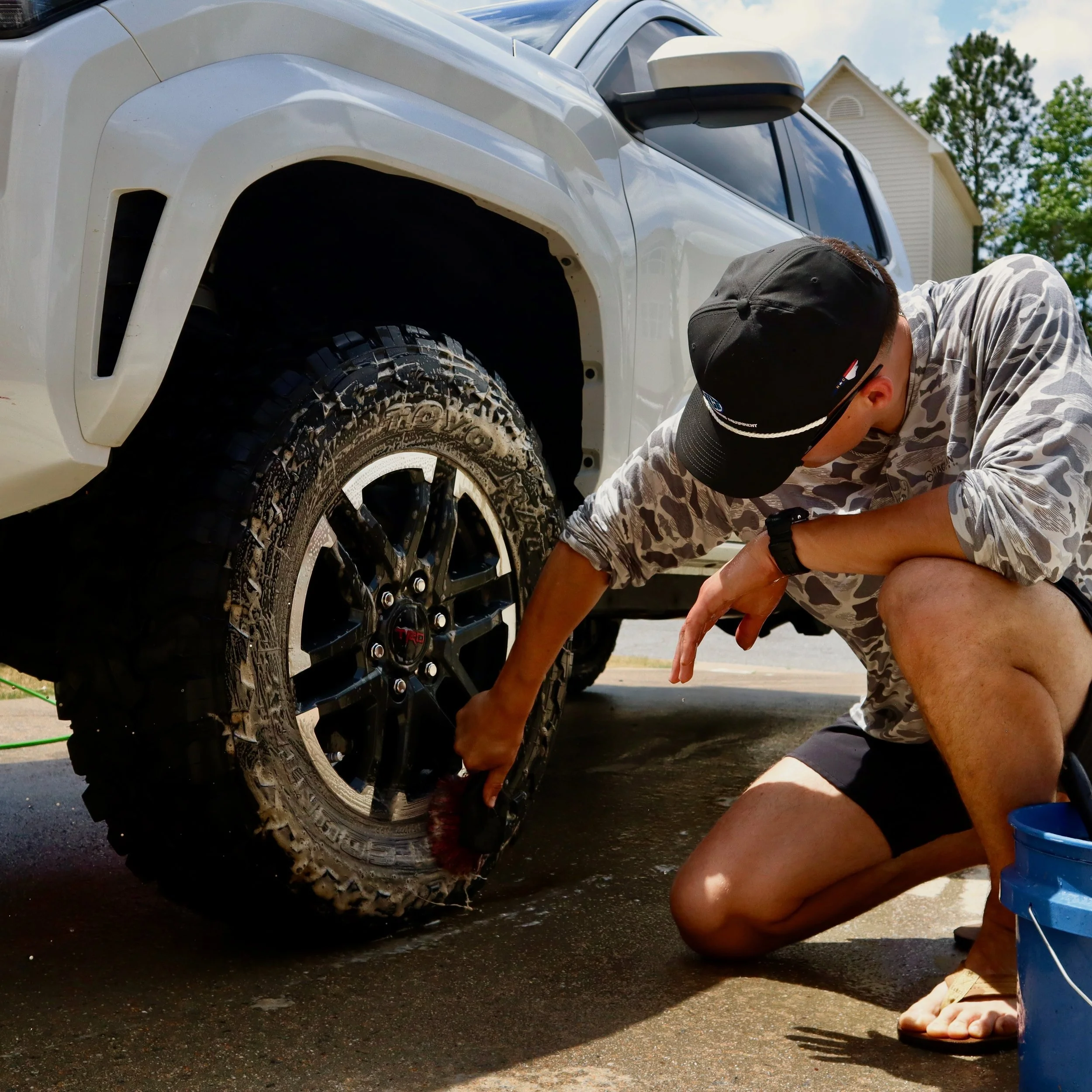 A person cleaning the dirt off a white pickup truck's tire with a brush outdoors during daytime.