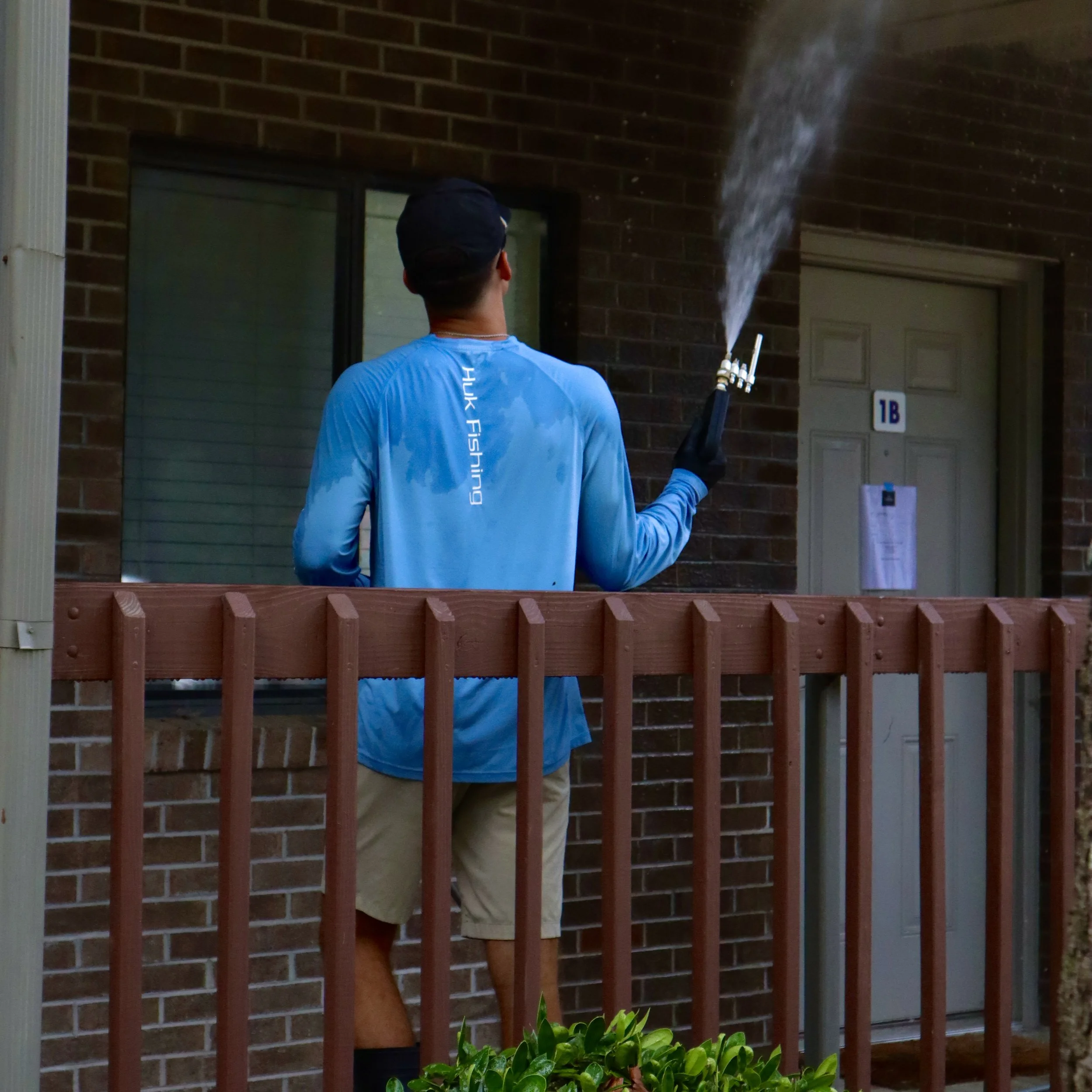 A man wearing a blue long sleeve shirt and khaki shorts, standing on a small porch, holding a fogger emitting fog or vapor near his shoulder, with his back to the camera.