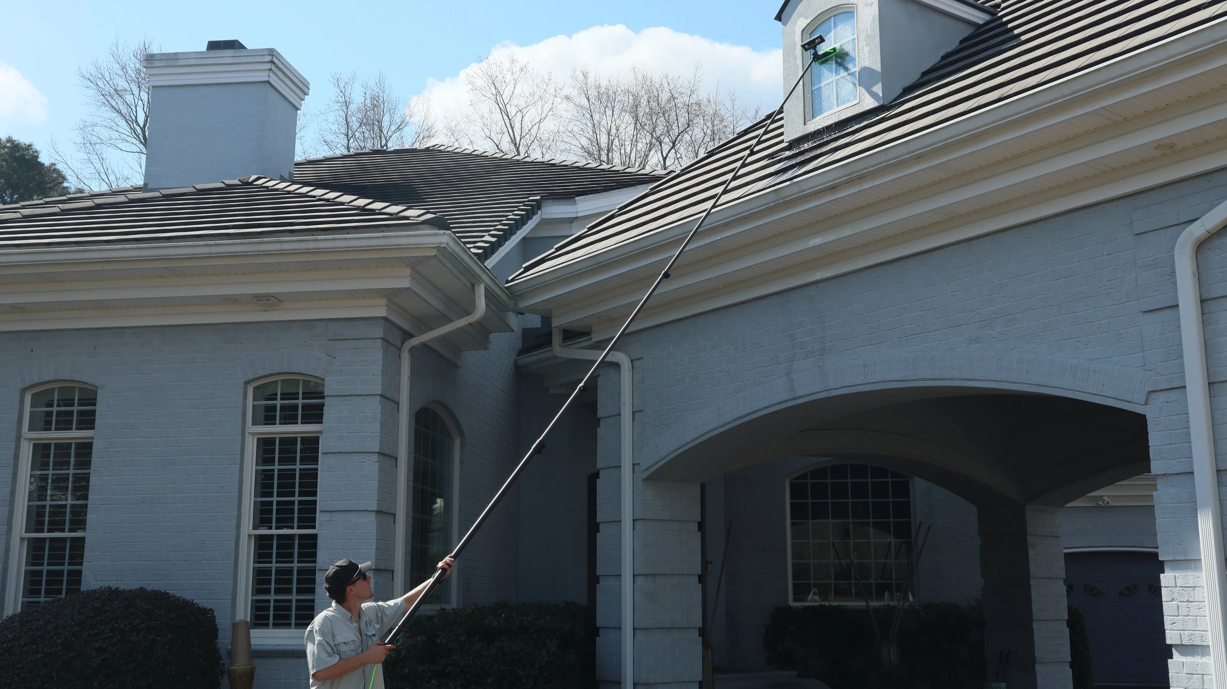 Person using an extendable pole to clean or inspect a high part of a house's roof, which has a white brick facade, large windows, and a garage, on a sunny day.