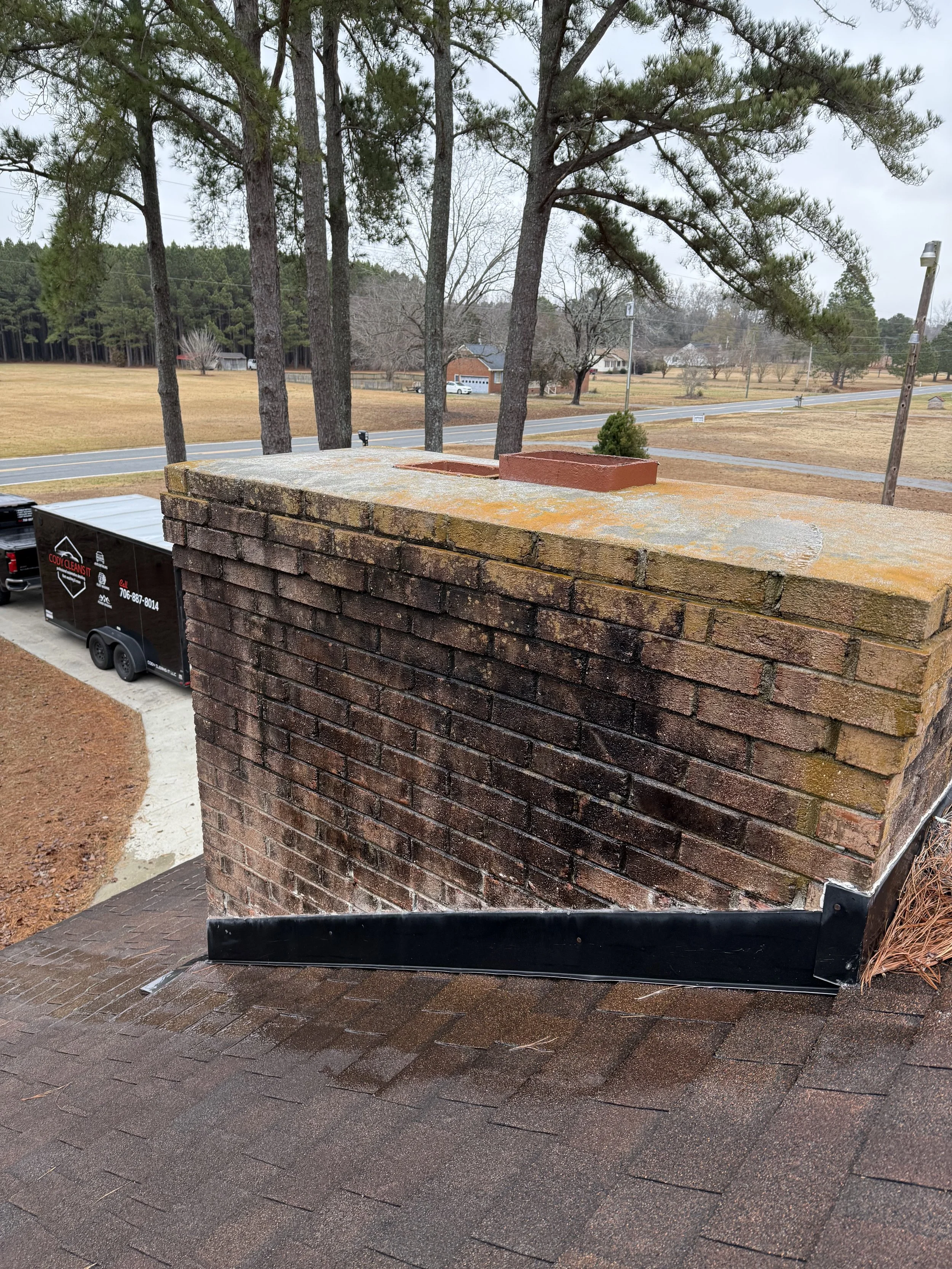 View of a brick chimney from a rooftop, with a red clay chimney cap visible on top. A black metal flashing at the base of the chimney and a dark brown shingled roof below. In the background, trees, a street, a trailer, and some houses are visible on a cloudy day.