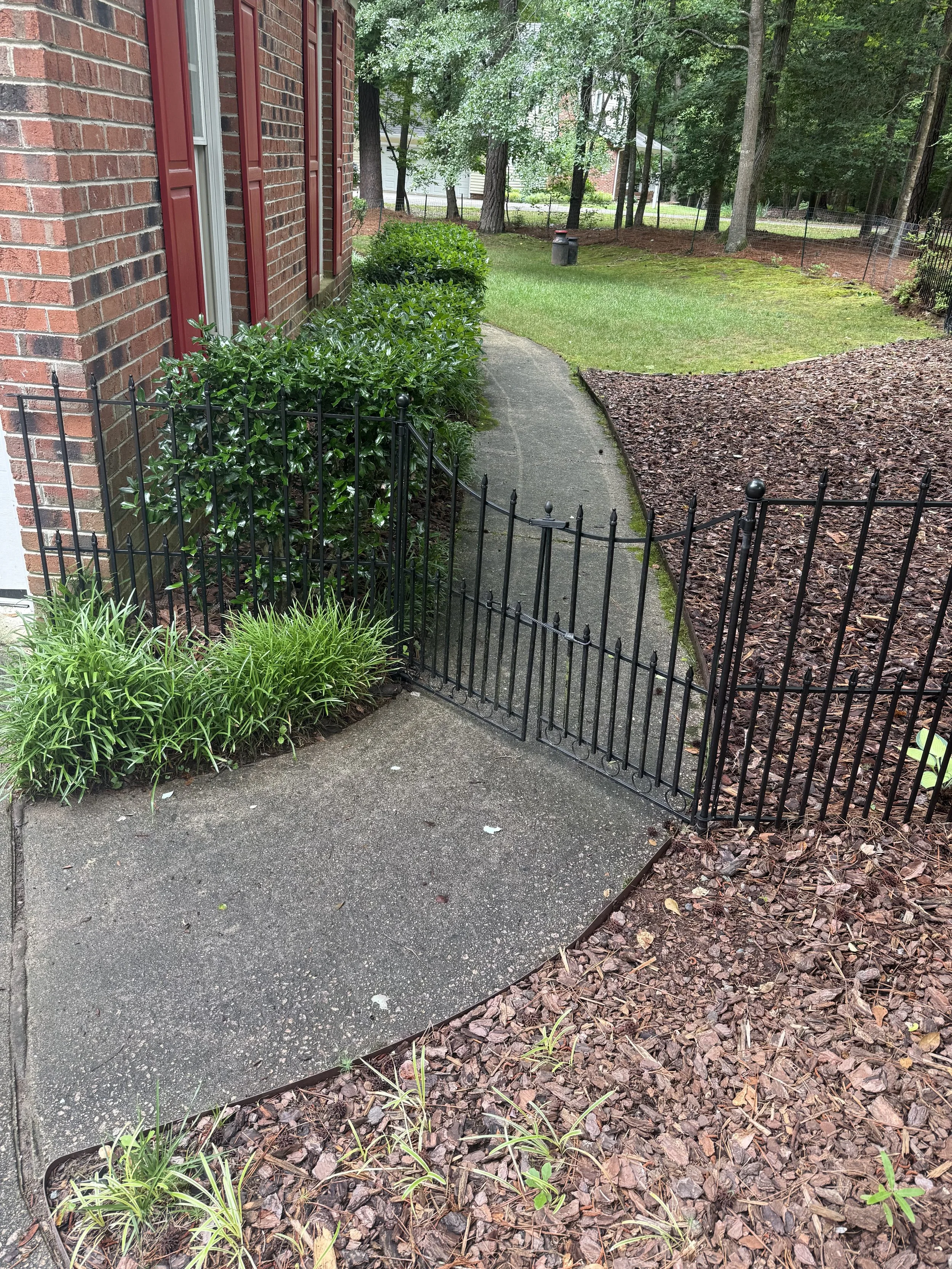 A concrete sidewalk curves alongside a brick house with red shutters. Green bushes are planted along the house, and a black metal fence surrounds part of the yard. The yard has green grass, trees, and a section with mulch. Trees and a residential area are visible in the background.