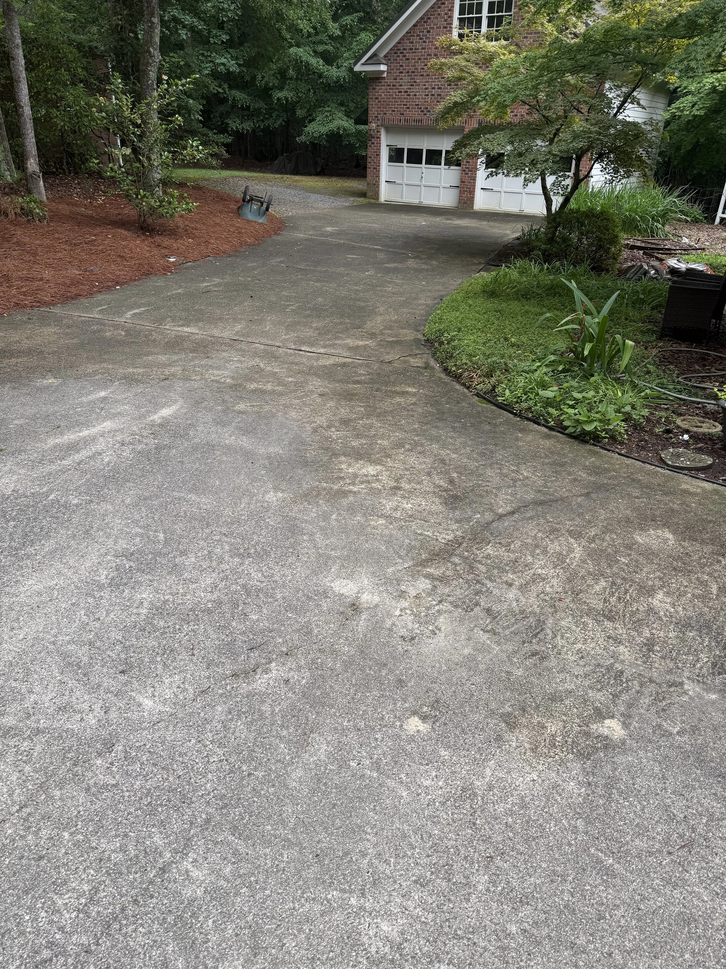 A concrete driveway in front of a brick house with a garage, surrounded by trees and greenery, with a wheelbarrow lying on its side.