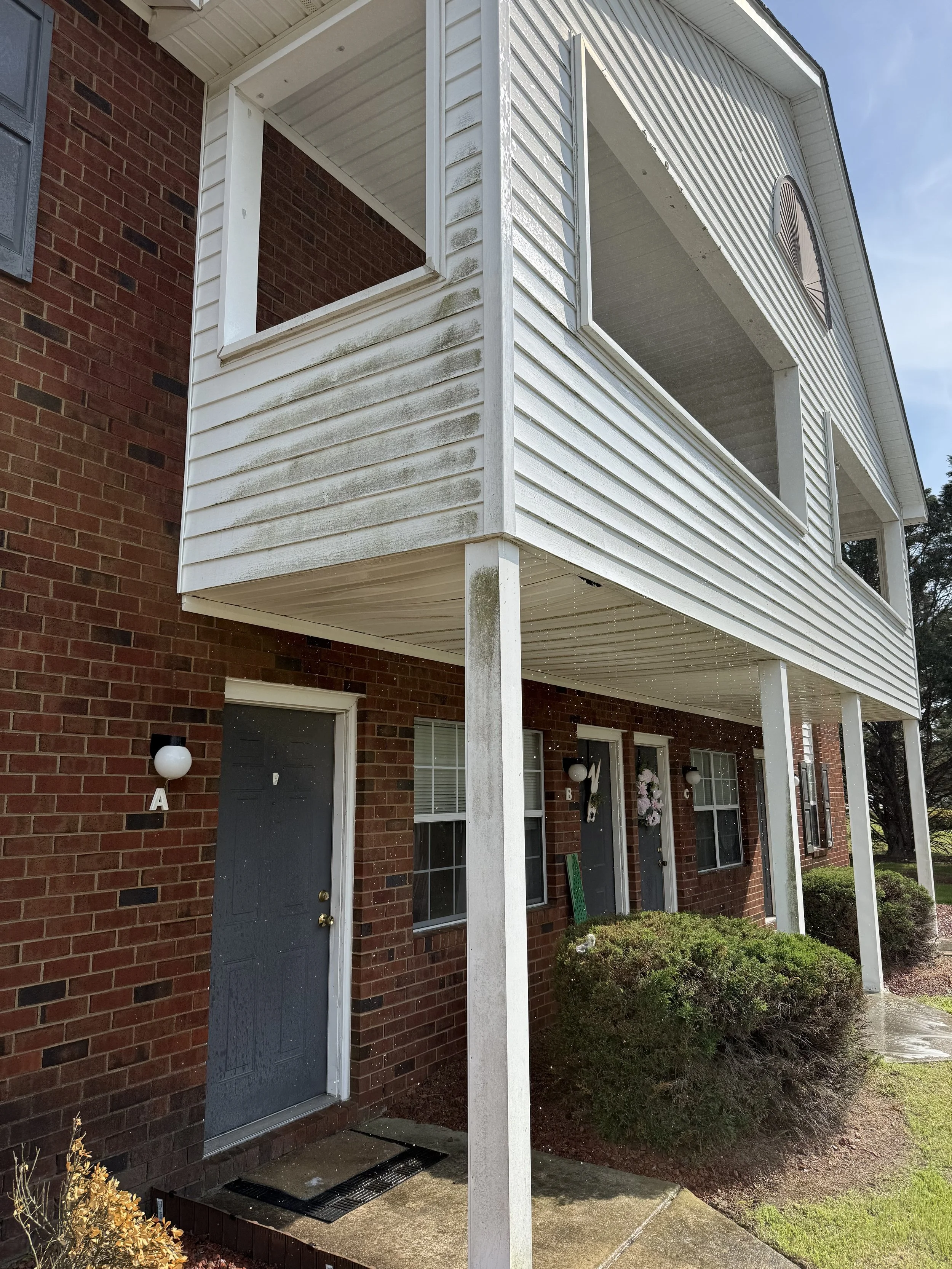 Apartment building with brick exterior and white siding on the balconies. The balconies are supported by white posts, with one balcony showing signs of dirt and moss. The building has multiple front doors, each with a window and a small outdoor wall lamp. The ground area in front of the doors has bushes and a concrete walkway.