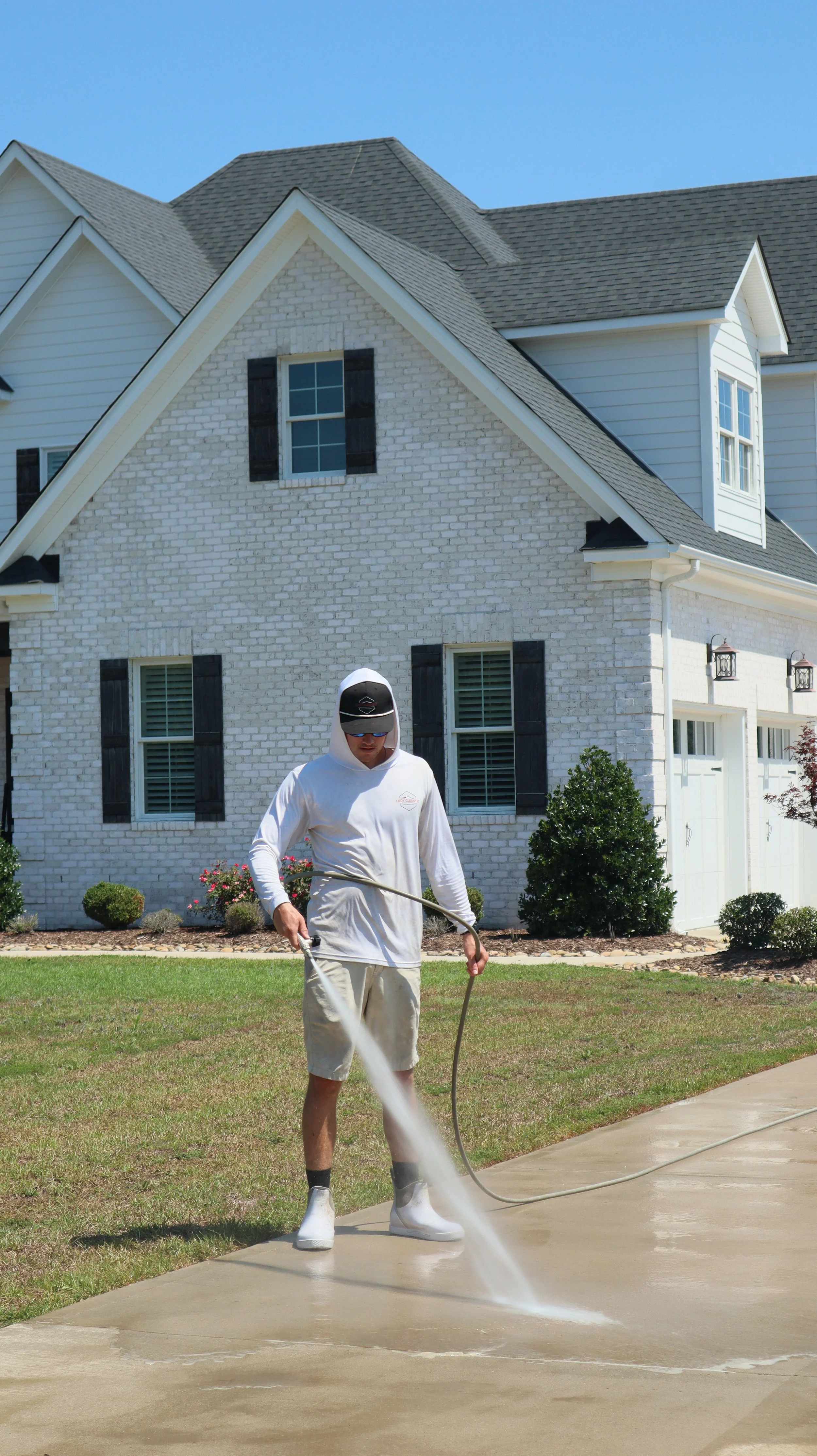 Person wearing white hoodie, beige shorts, black socks, and white shoes spraying concrete on a driveway in front of a white brick house with black shutters.