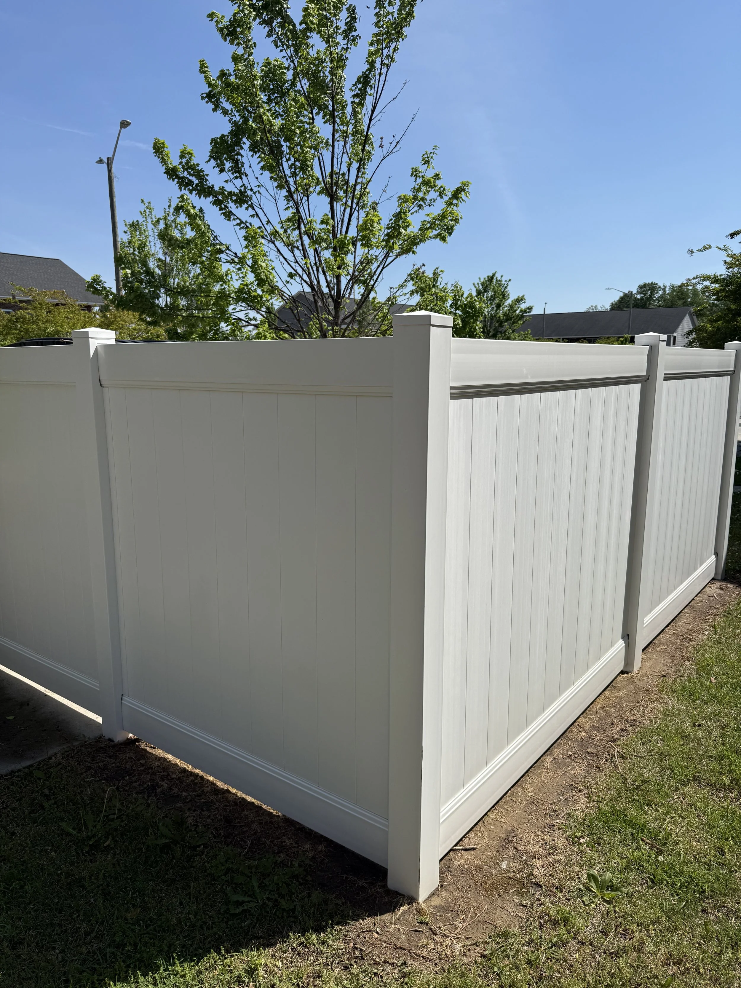 A white privacy fence surrounds a yard with green grass and trees, under a clear blue sky with a single streetlight visible in the background.