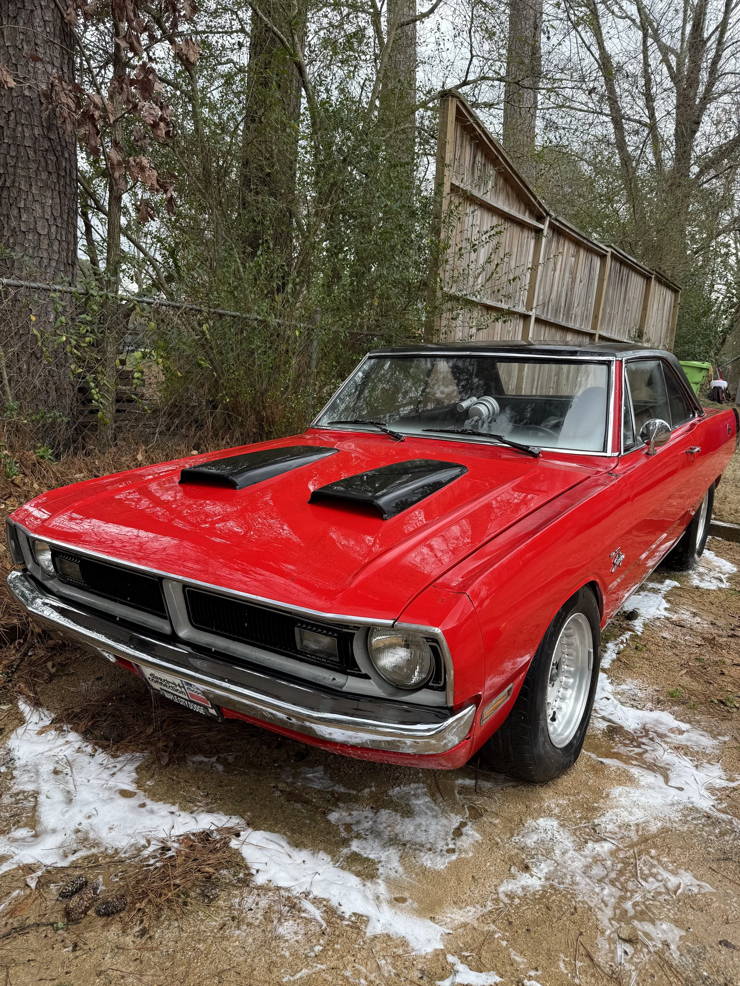 A red vintage muscle car with black racing stripes parked on a dirt ground with patches of snow, surrounded by trees and a wooden fence in the background.