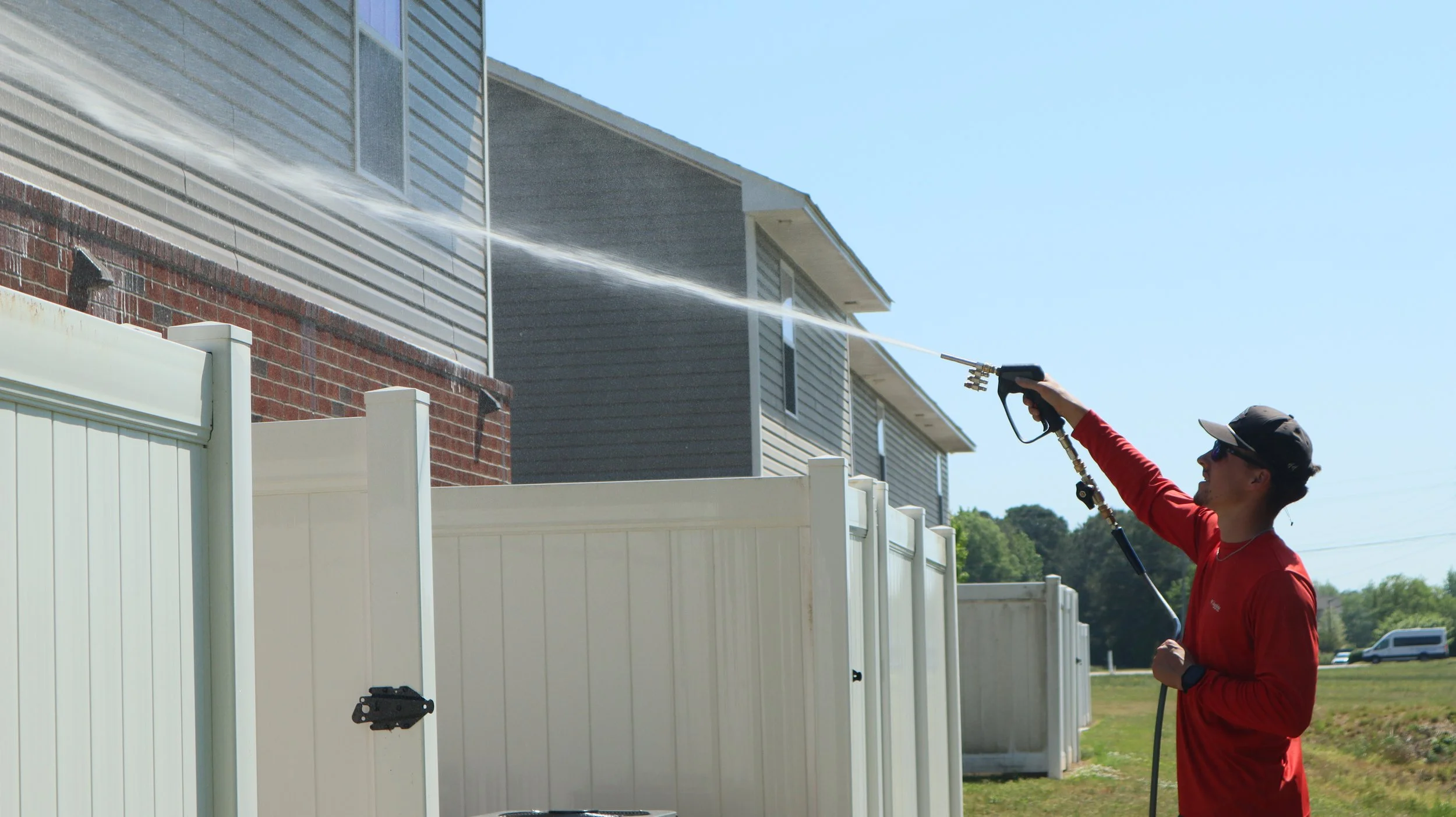 A person wearing a red long sleeve shirt, sunglasses, and a cap is pressure washing a white fence outdoors on a sunny day. Behind them are houses with gray and brick exteriors, and there is a grassy area with trees in the background.