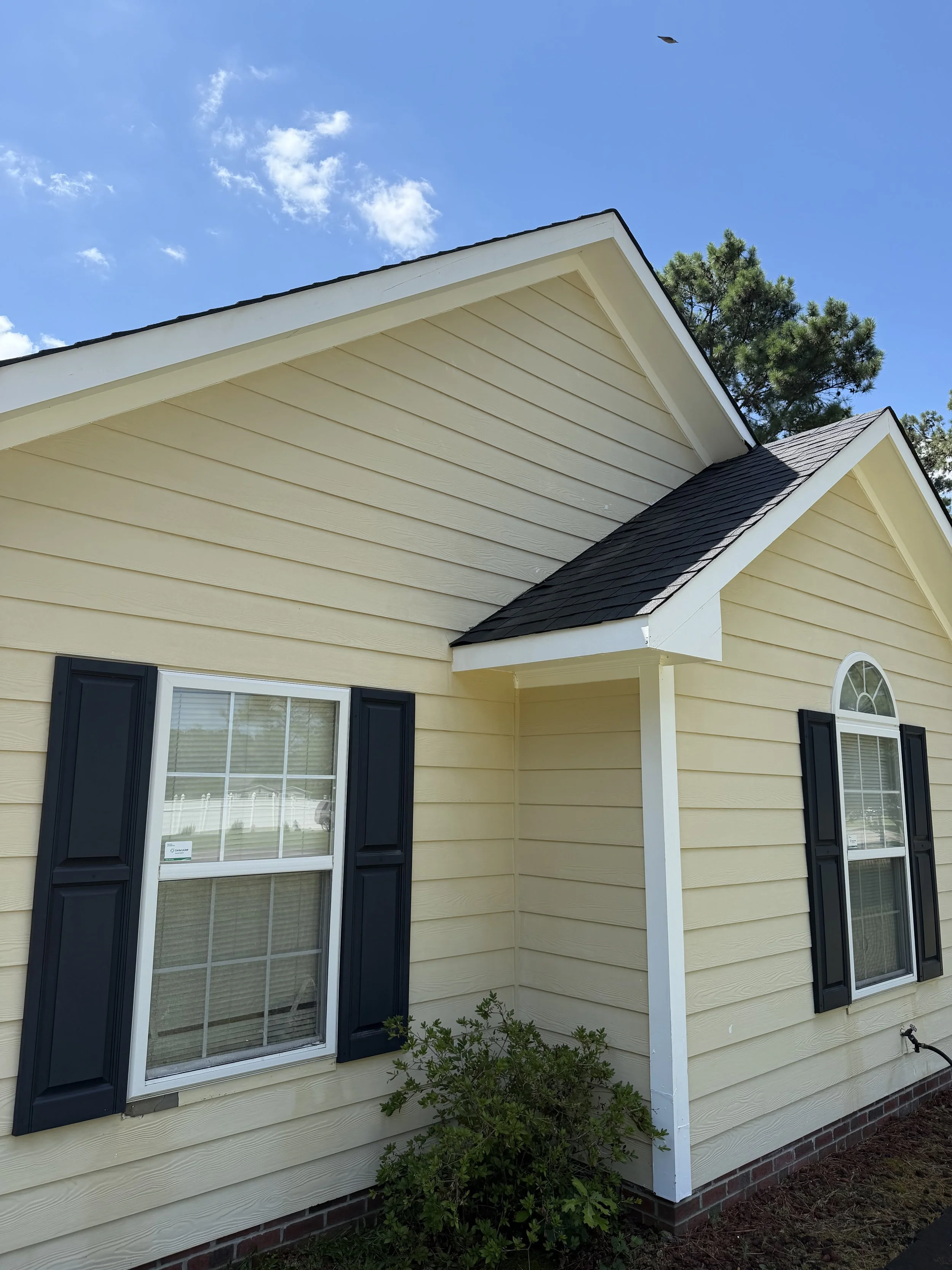 Exterior view of a yellow house with white trim, black shutters, and large window with grid pattern, showing a small shrub in front and a clear blue sky with some clouds.
