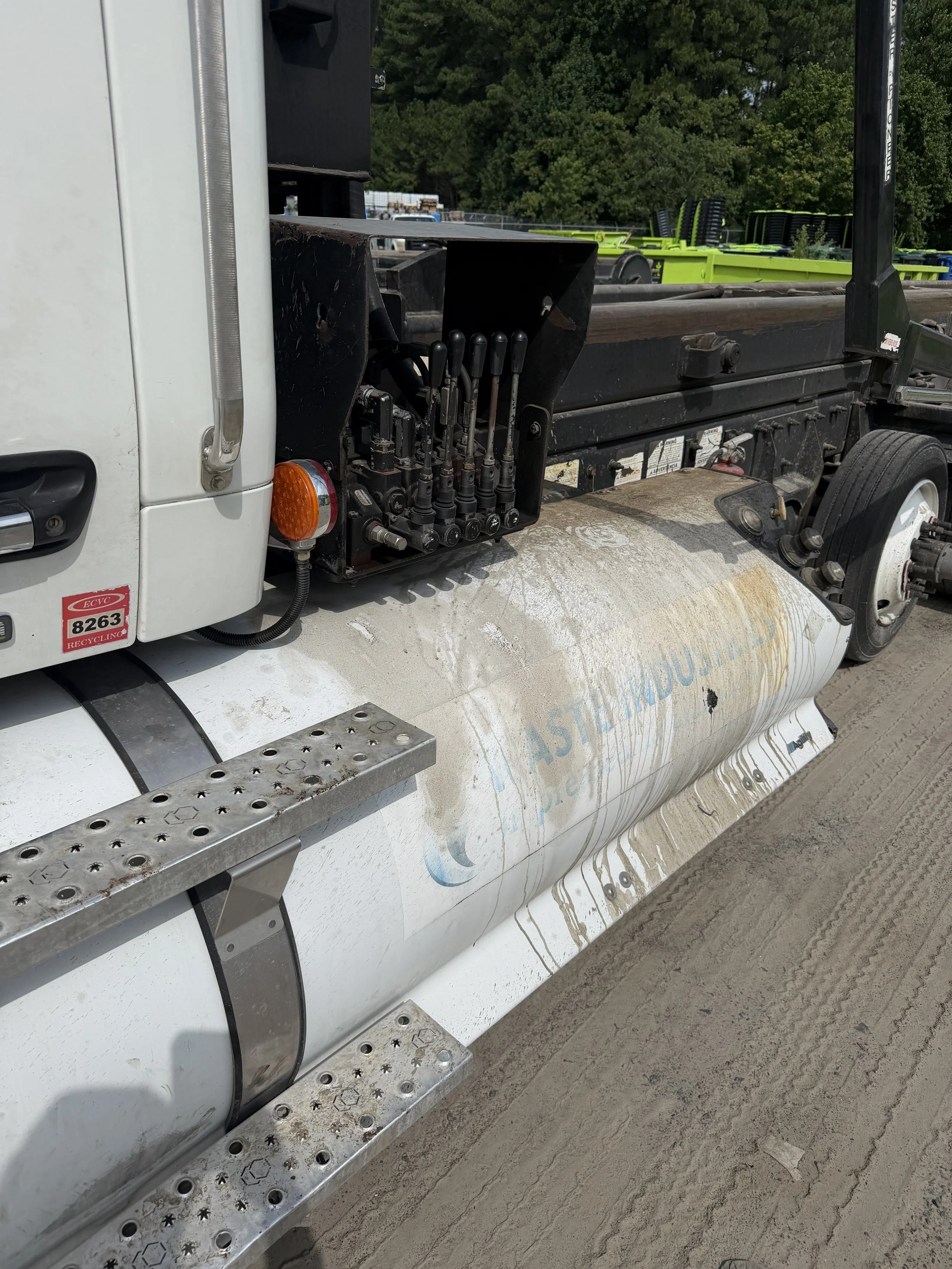 Close-up of a truck's side panel showing a row of levers and a metal step with perforations, with soil and some dirt on the truck surface, and green trees in the background.