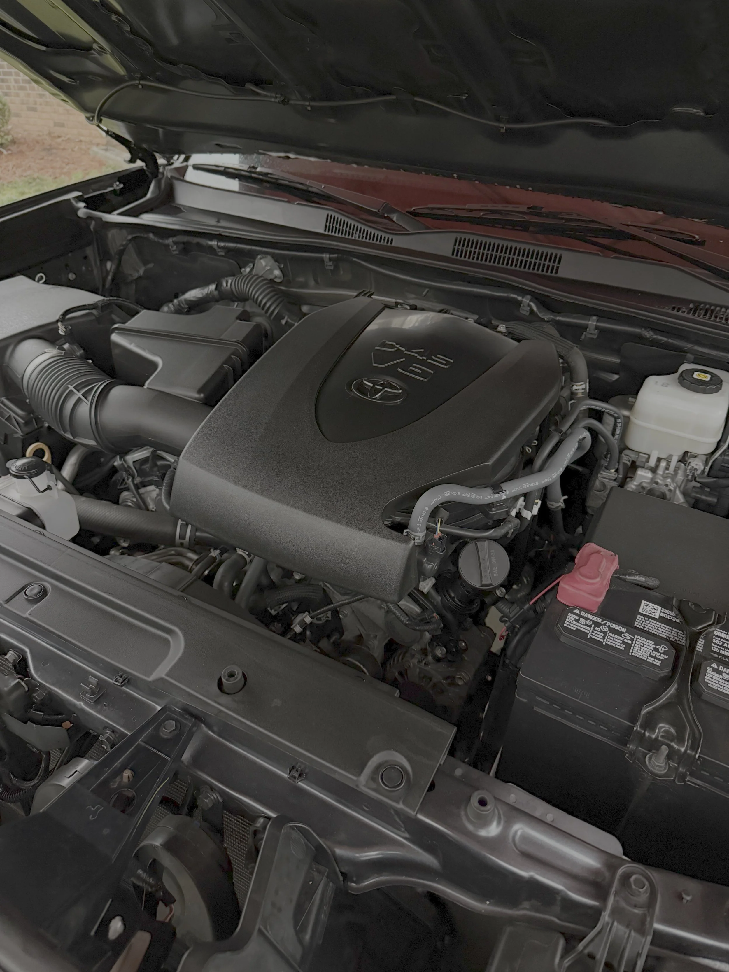 View of a car engine bay, showing various components including hoses, filters, and the engine block, with the hood open.