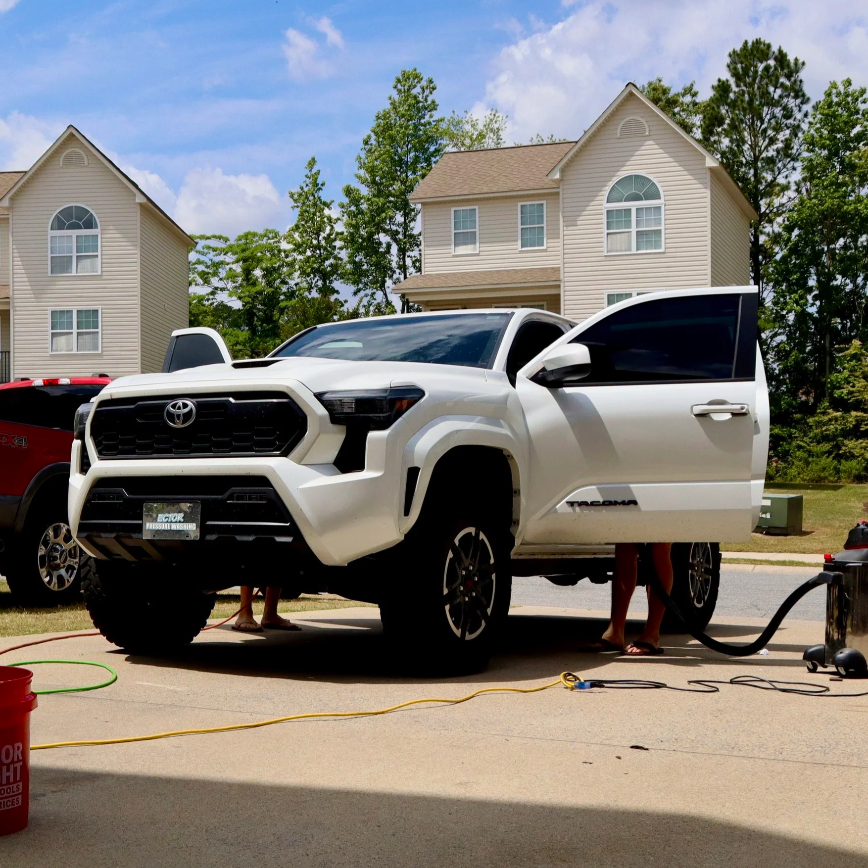 People washing a white Toyota Tacoma pickup truck in a driveway using pressure washers, with a suburban house and trees in the background.