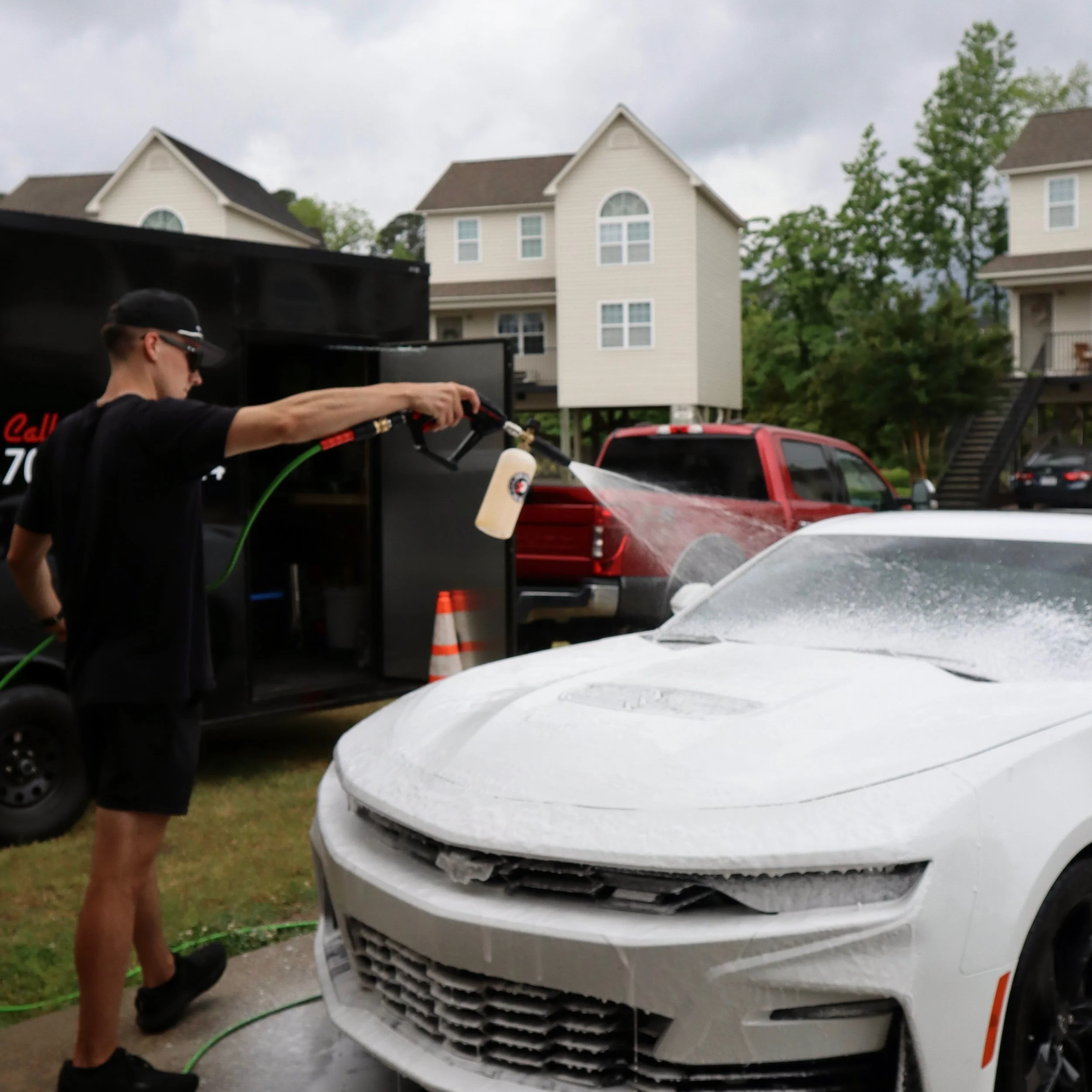 A person wearing black shorts, t-shirt, and cap washing a white sports car with foam using a pressure washer outdoors, with houses and trees in the background.