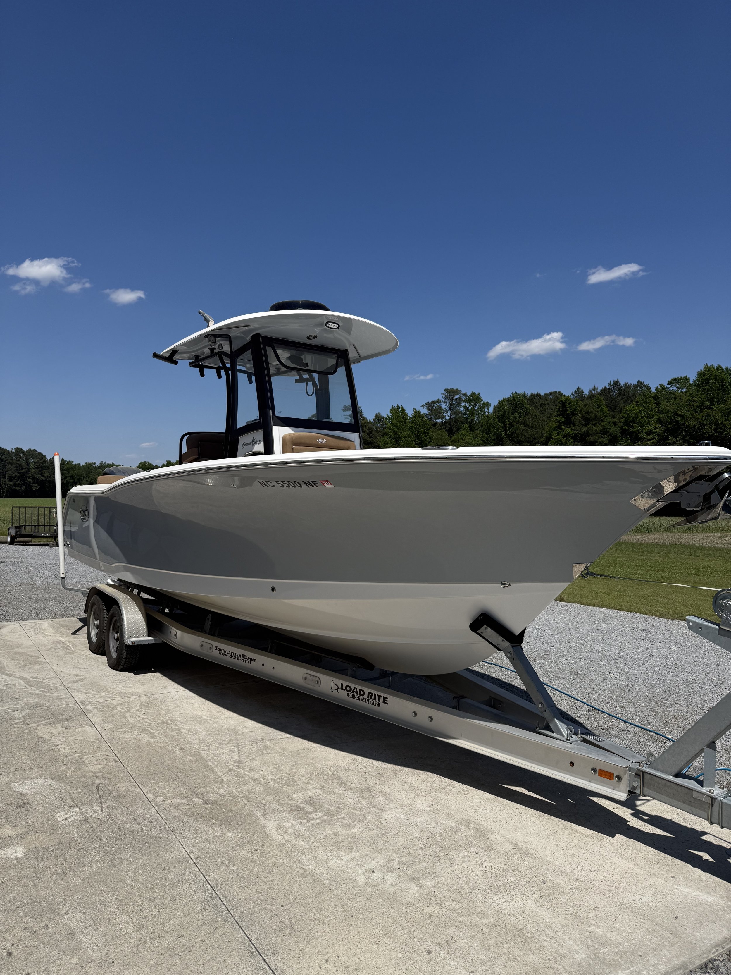 A white boat on a trailer parked on a concrete surface with trees and a blue sky with a few clouds in the background.