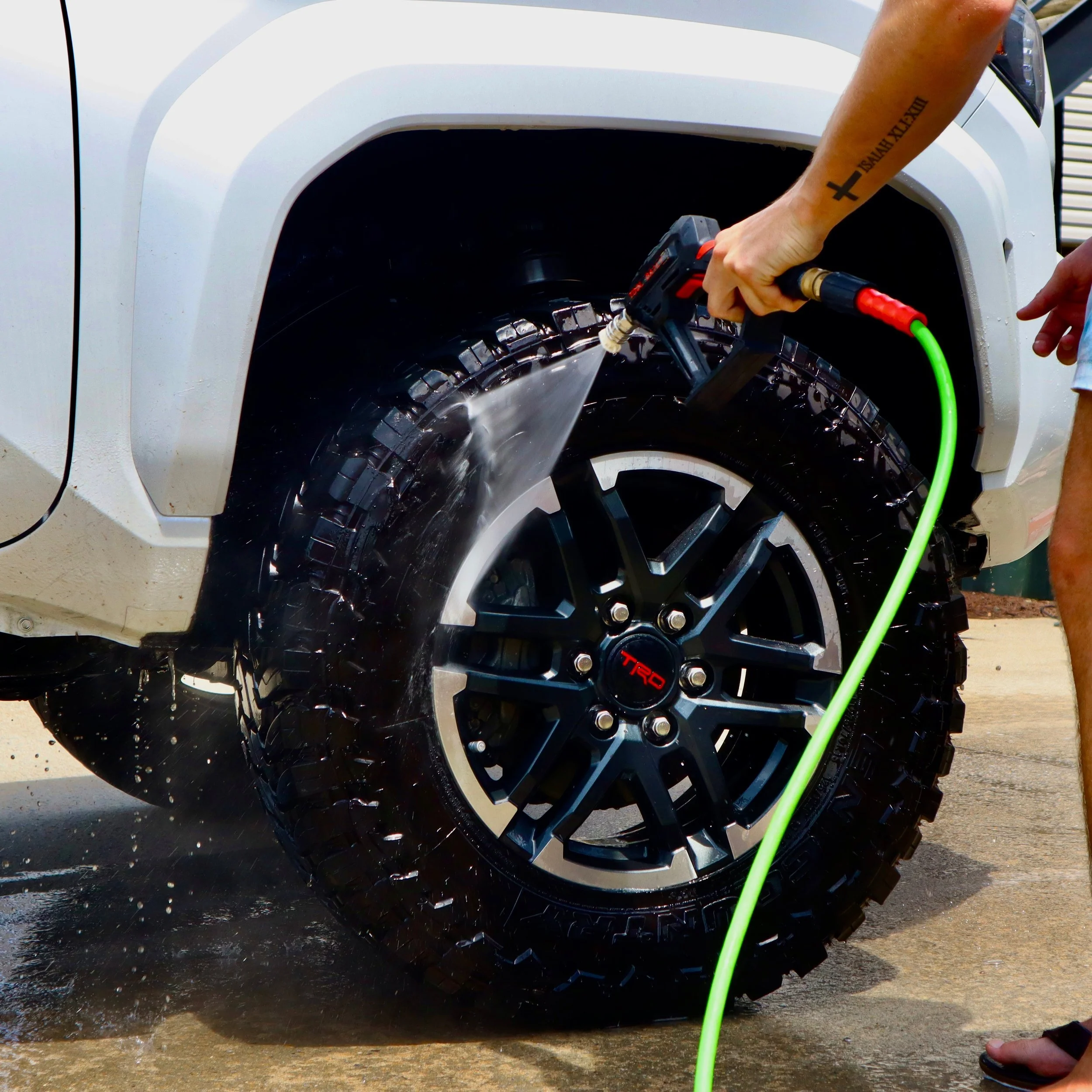 Person washing a black tire of a white vehicle using a pressure washer, with a yellow garden hose on the ground.