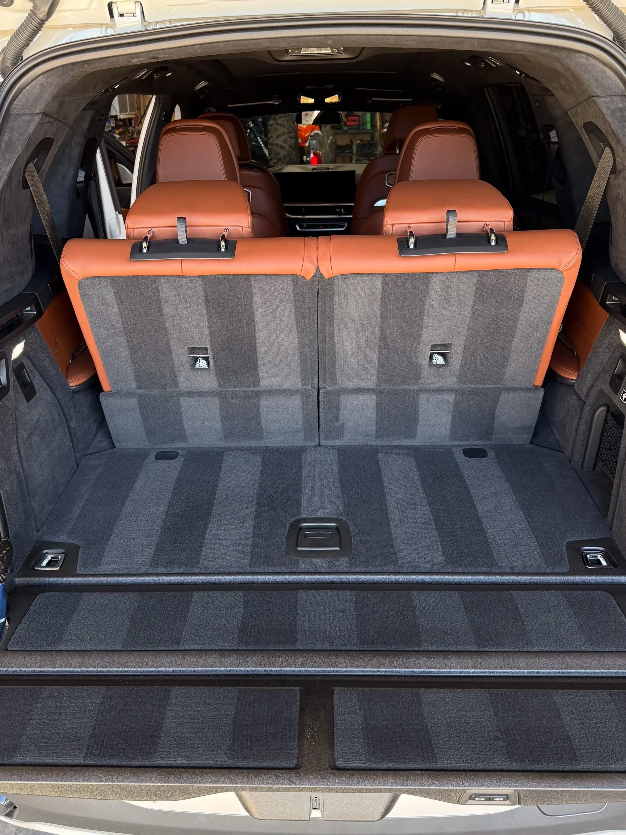 The interior of an SUV with the third-row seats folded down, showing cargo space with a black carpeted floor and brown leather seats.