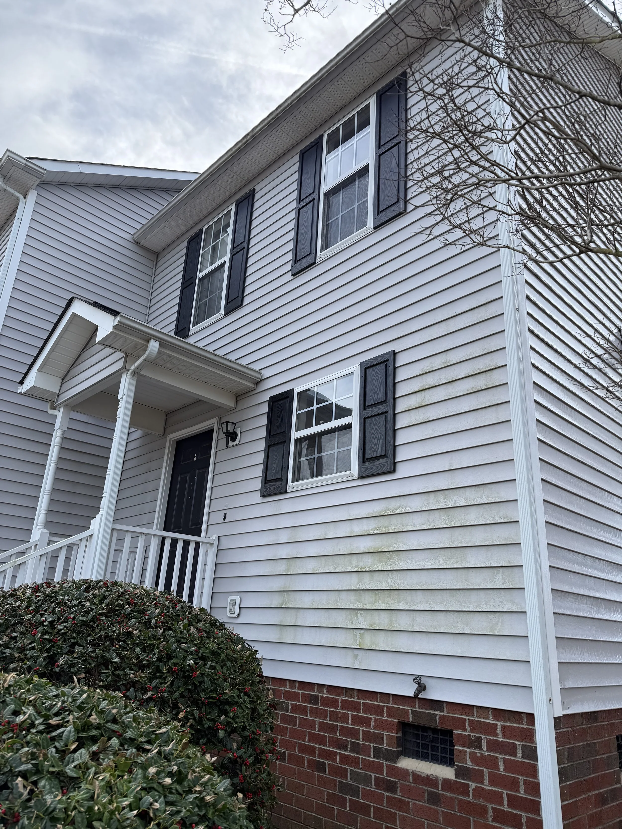 Exterior view of a two-story house with white vinyl siding, black window shutters, and a brick foundation. There are trees with bare branches and a bush with red berries in the foreground.