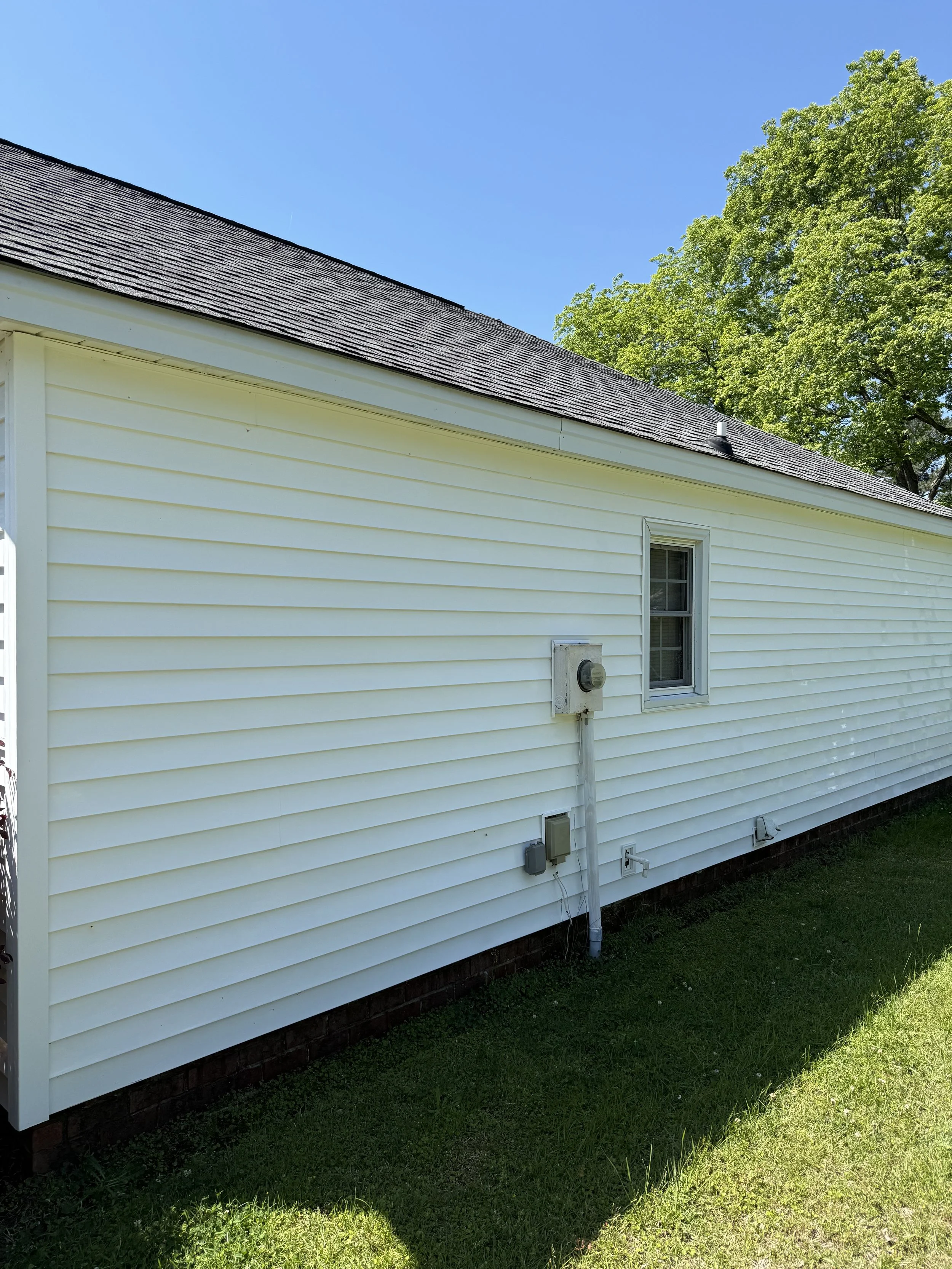 Side view of a house with white vinyl siding, a small window, electrical boxes, and a dark shingled roof under a blue sky with green trees in the background.