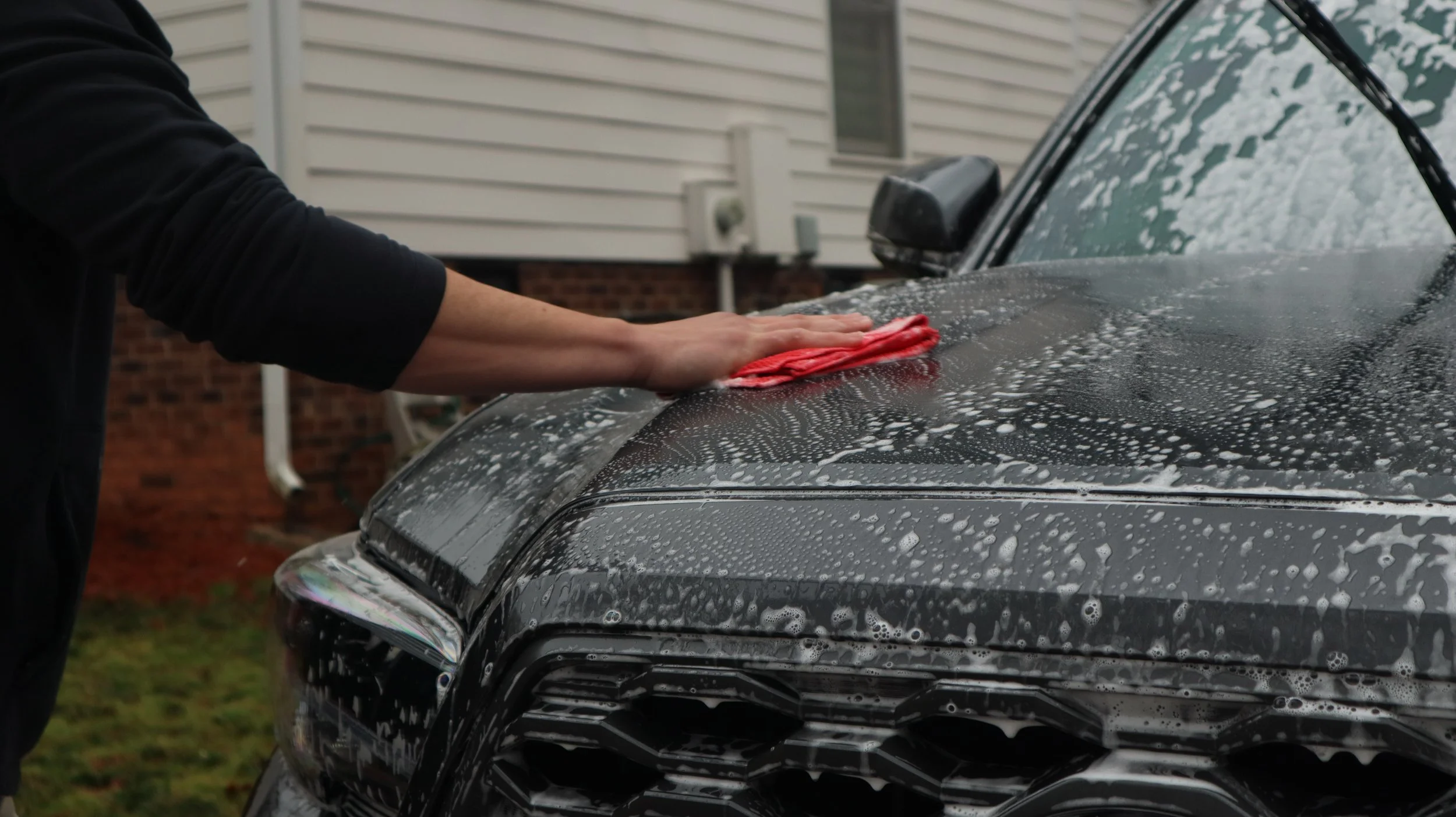 Person washing a black car with a red cloth, water and soap suds on the vehicle, outdoor residential setting.