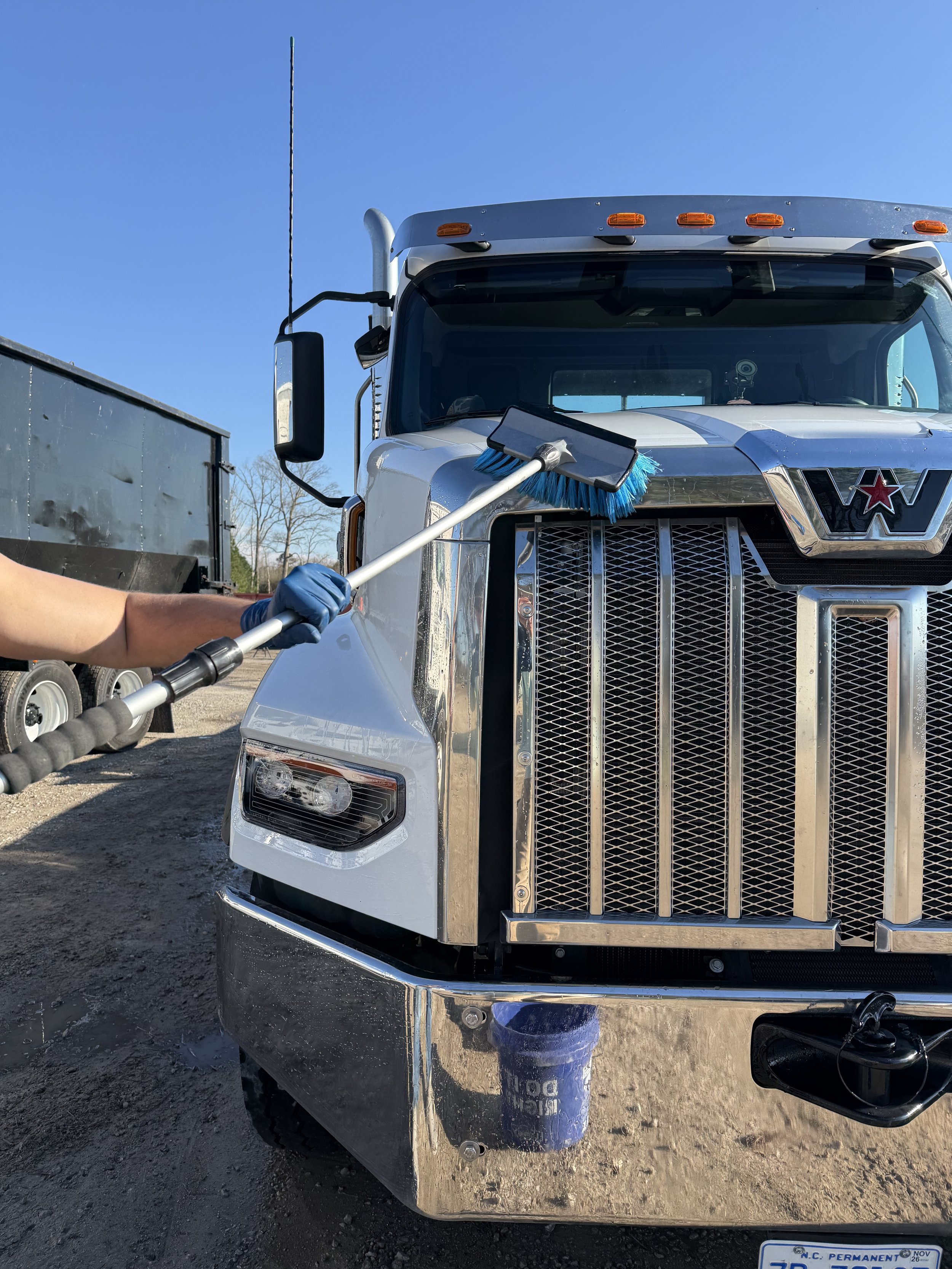 Front of a white semi-truck with a street sweeper brush attached to the front, on a dirt lot with a blue sky.