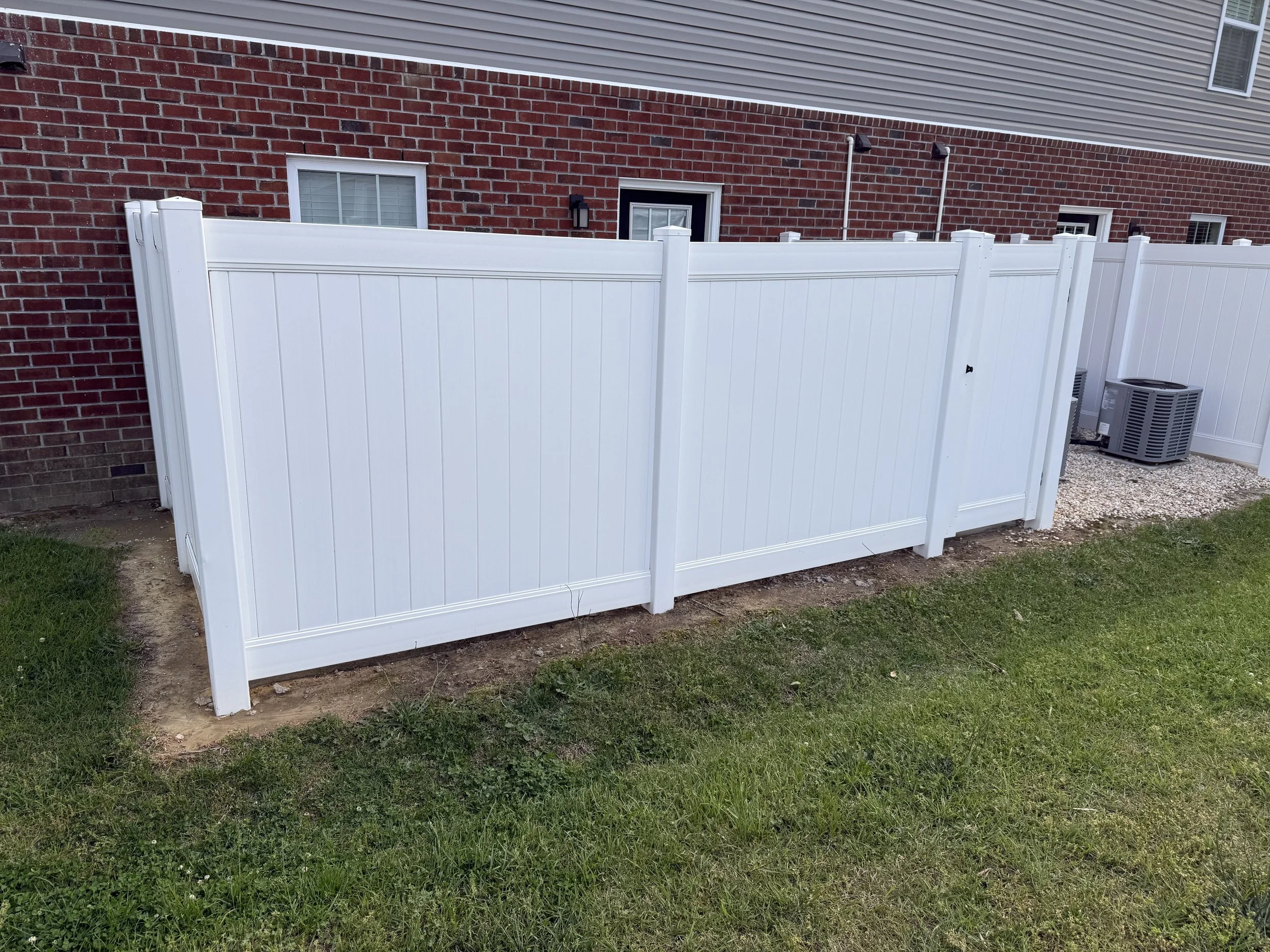 A white privacy fence with a house in the background, red brick wall, grey siding, multiple windows, two outdoor wall lights, and an air conditioning unit on a gravel pad.