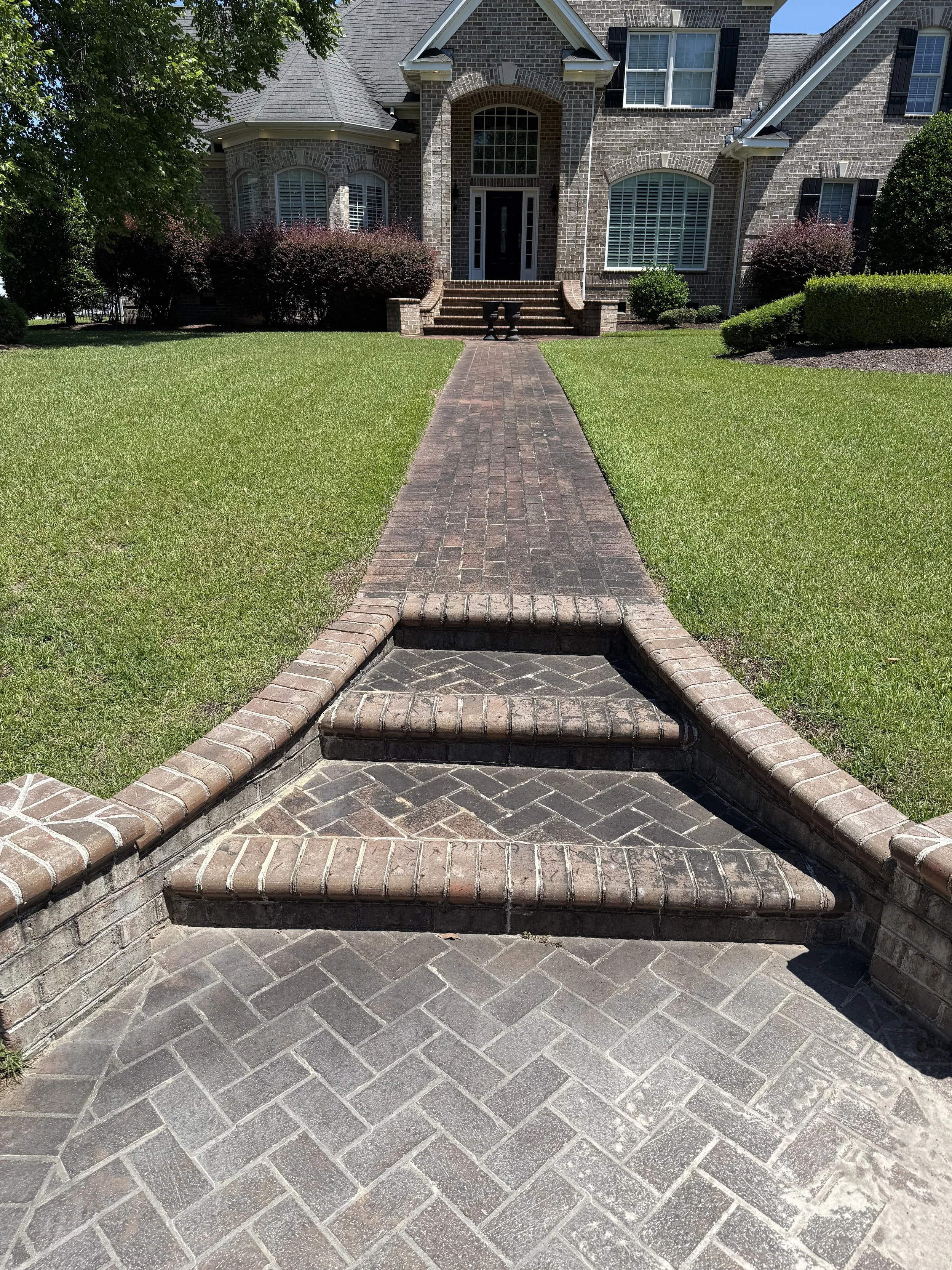 A brick sidewalk leading up to a house with a brick front door, brick stairs, and multiple windows with shutters, surrounded by a well-manicured lawn and bushes.