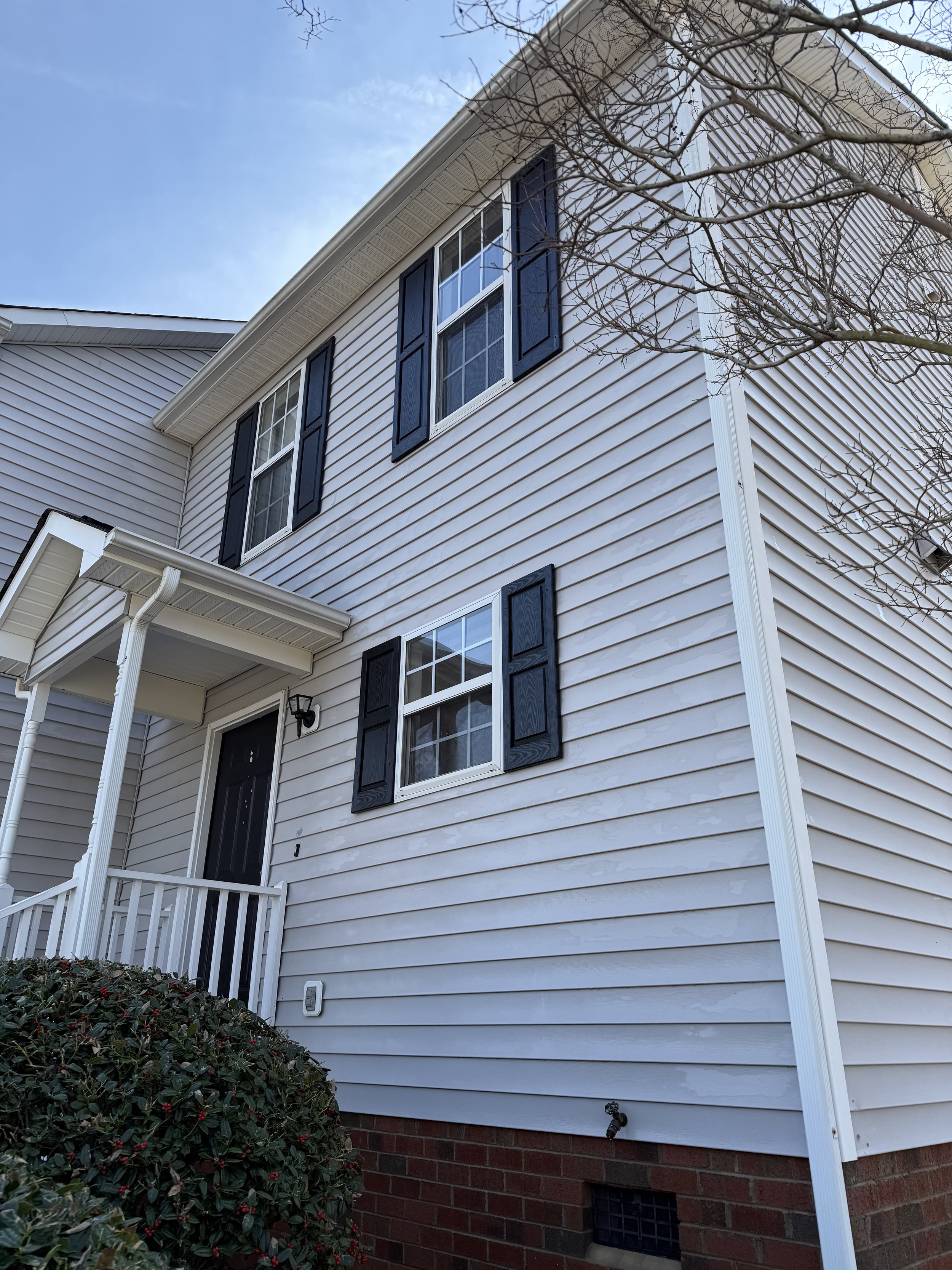 A two-story house with white siding and black shutters, featuring a front porch with white railings, a small outdoor light, and surrounded by leafless tree branches and green bushes with red berries. The lower part of the house has a brick foundation, and a small vent is visible near the ground.