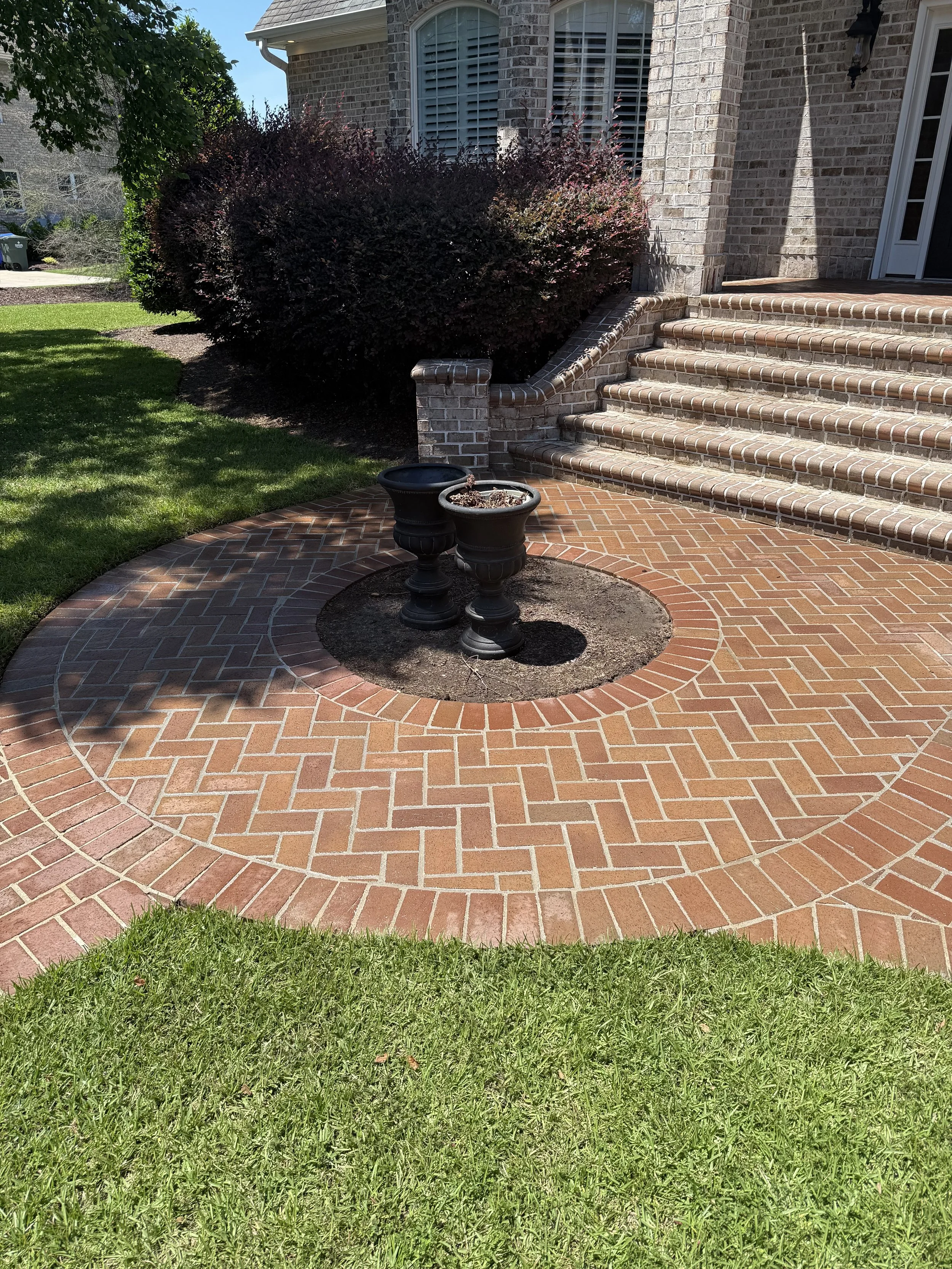 Photo of the front steps and porch of a brick house with two black planters in a circular brick-paved area, situated next to a lawn with green grass, and a bush with dark red leaves near the stairs.
