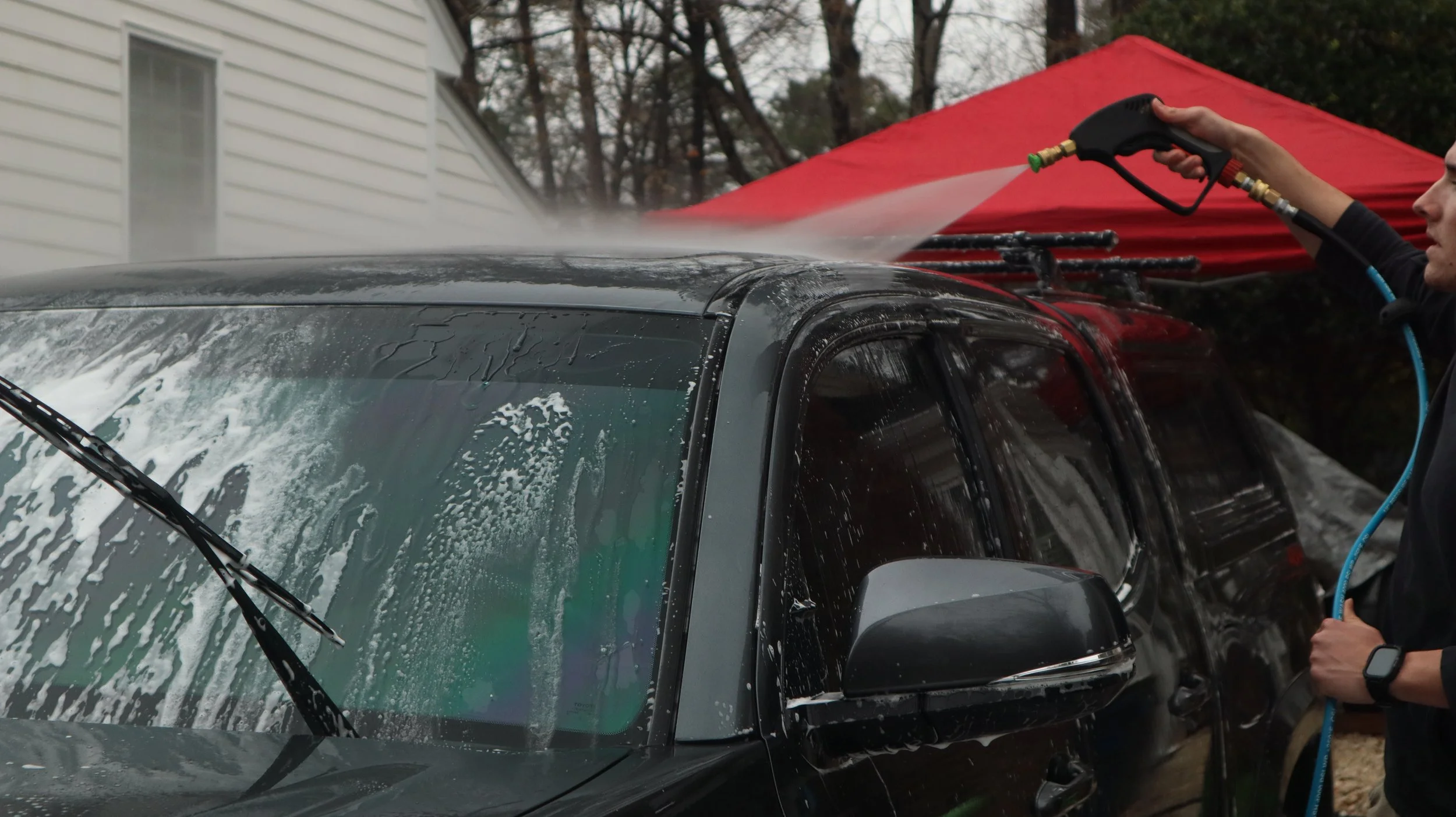 Person pressure washing a black car outdoors on a cloudy day, with a red canopy and trees in the background.