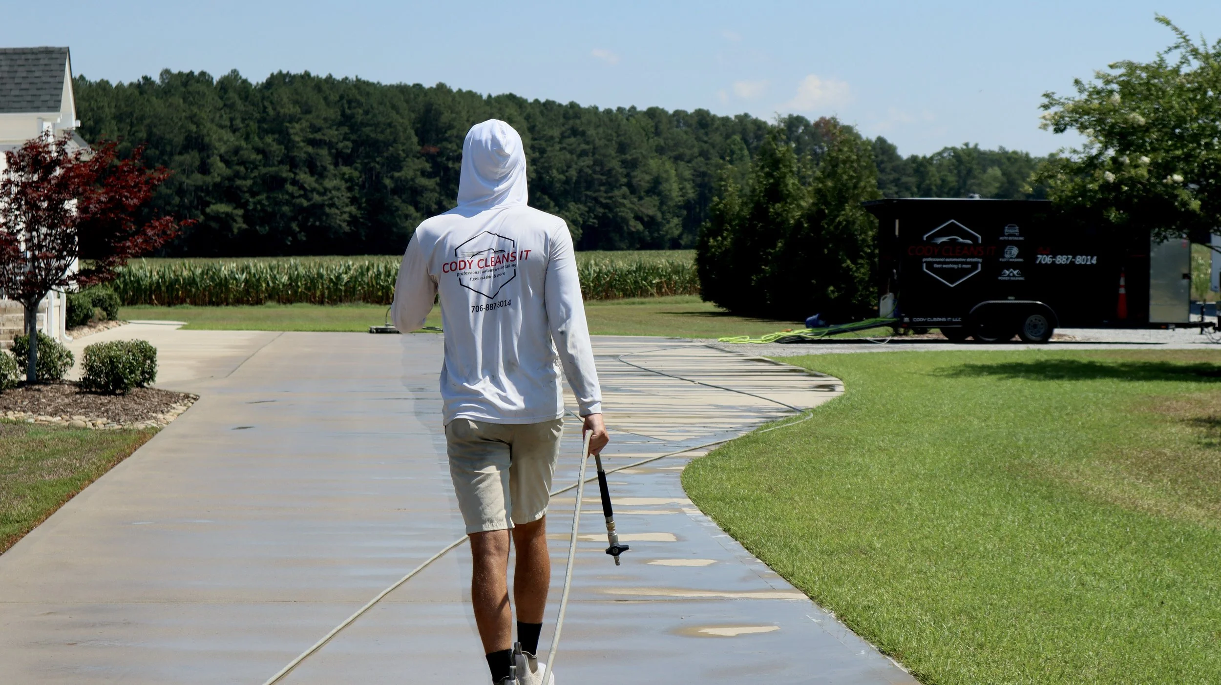Person in white hoodie walking on a sidewalk with a cane, near a trailer and a green field with trees in the background.