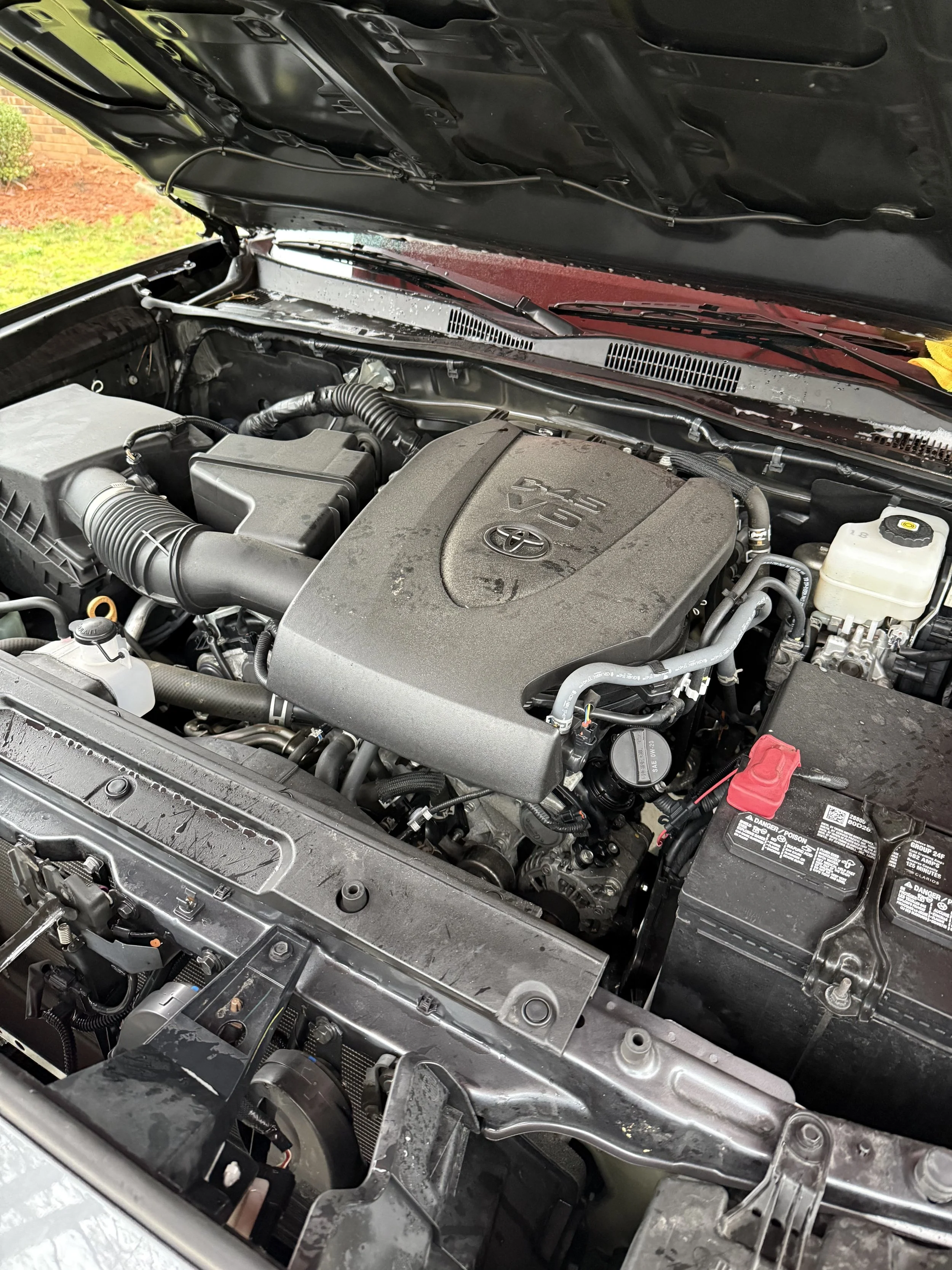 The image shows a close-up view of a car engine with the hood open, revealing various engine components, hoses, and parts.