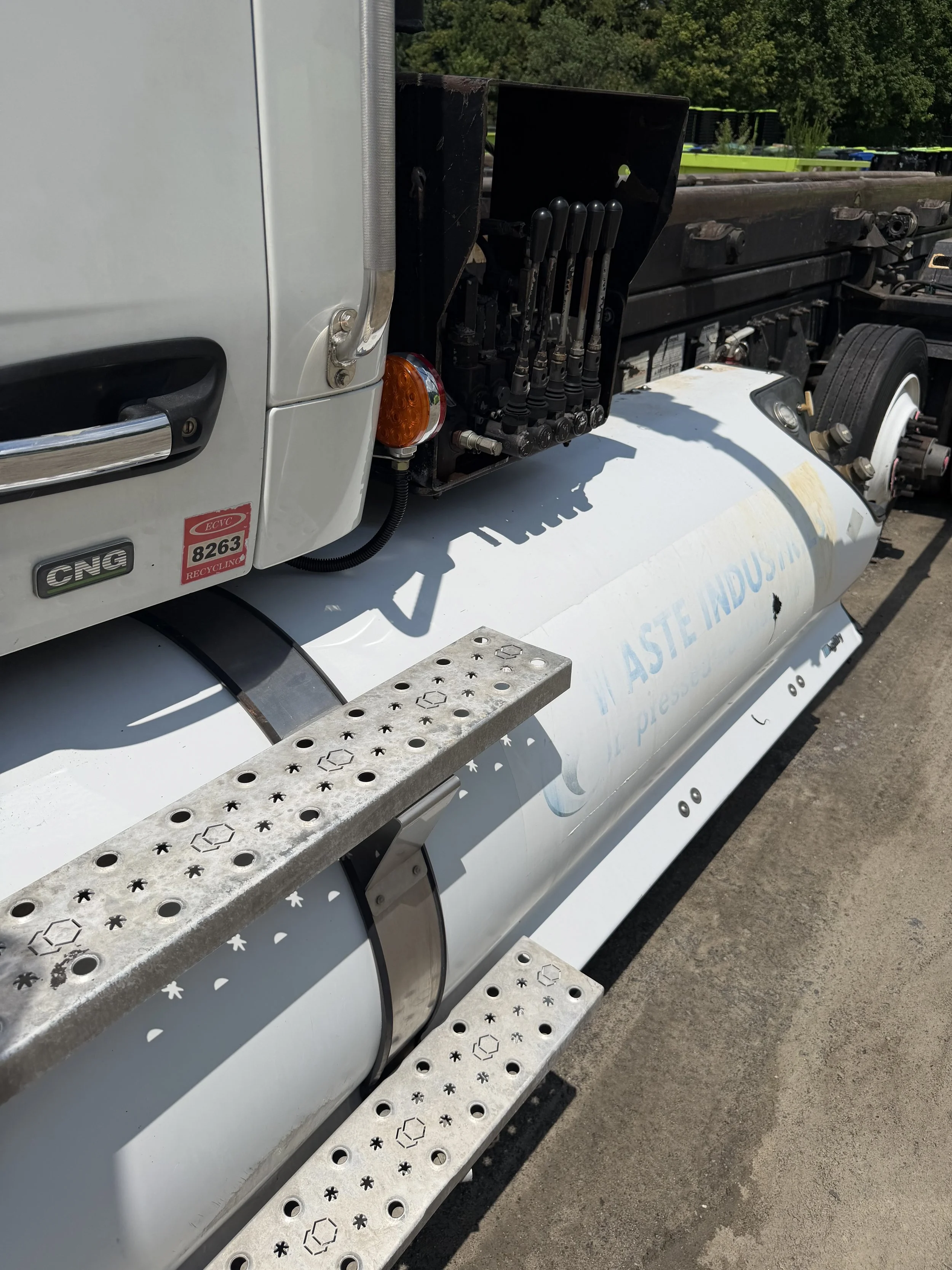 Close-up of a truck's side, showing a set of levers, a step, and some decals indicating CNG fuel and recycling.