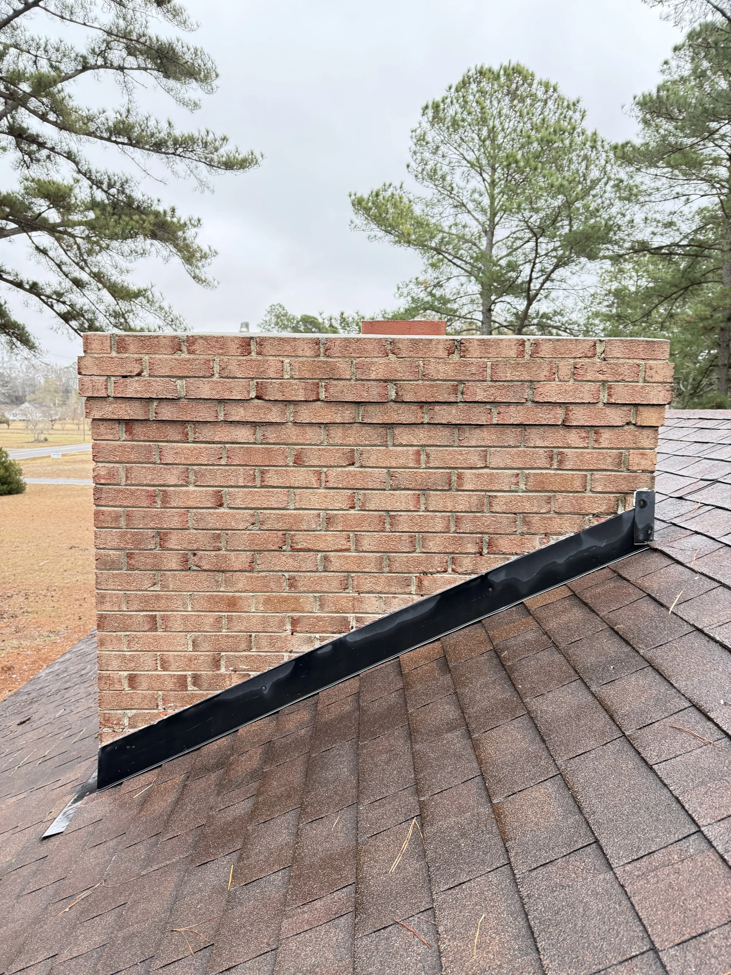 Close-up of a red brick chimney on a residential roof, with brown asphalt shingles and a black metal flashing at the base of the chimney, against a cloudy sky and pine trees in the background.