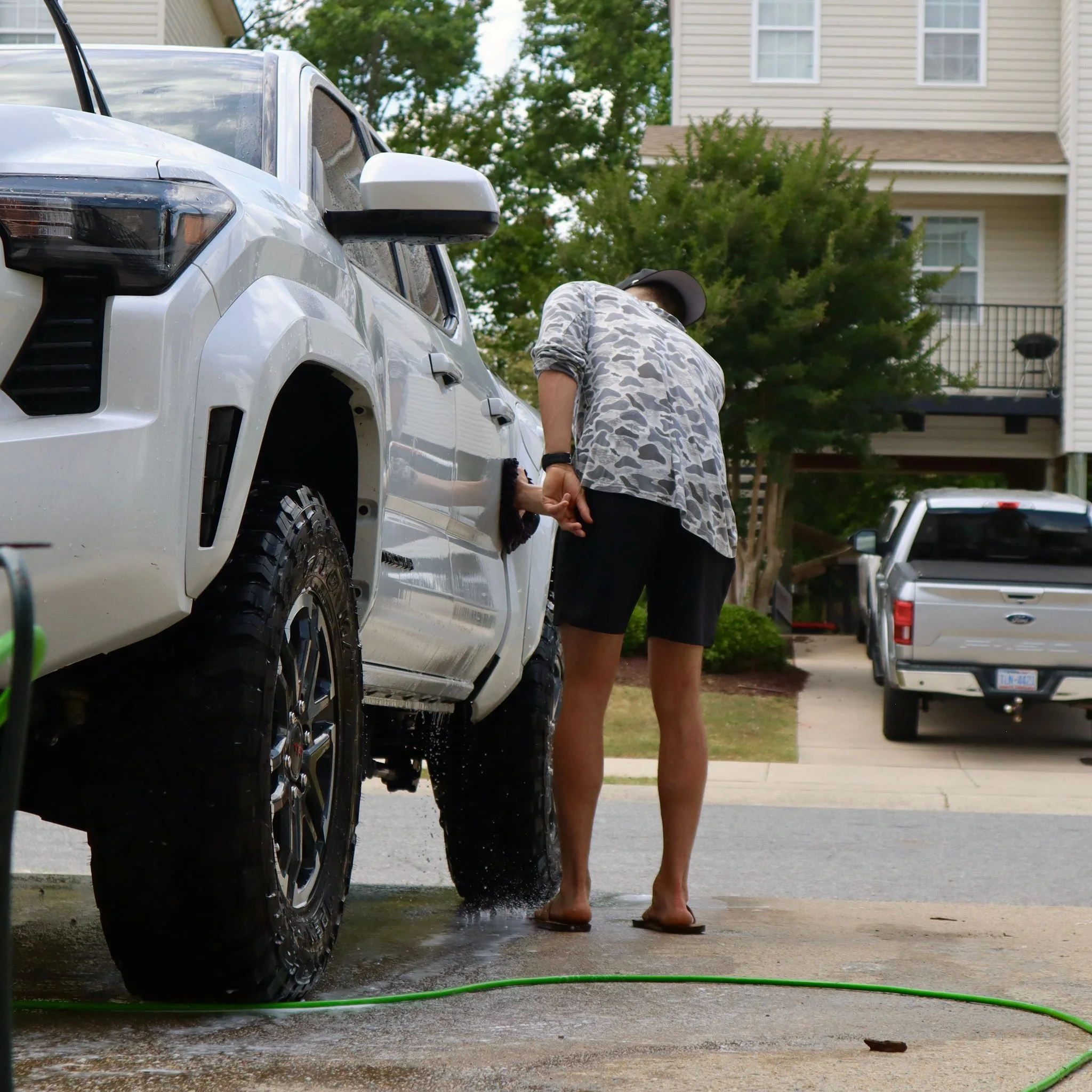 Person washing a white pickup truck outside on a driveway, with a hose on the ground.