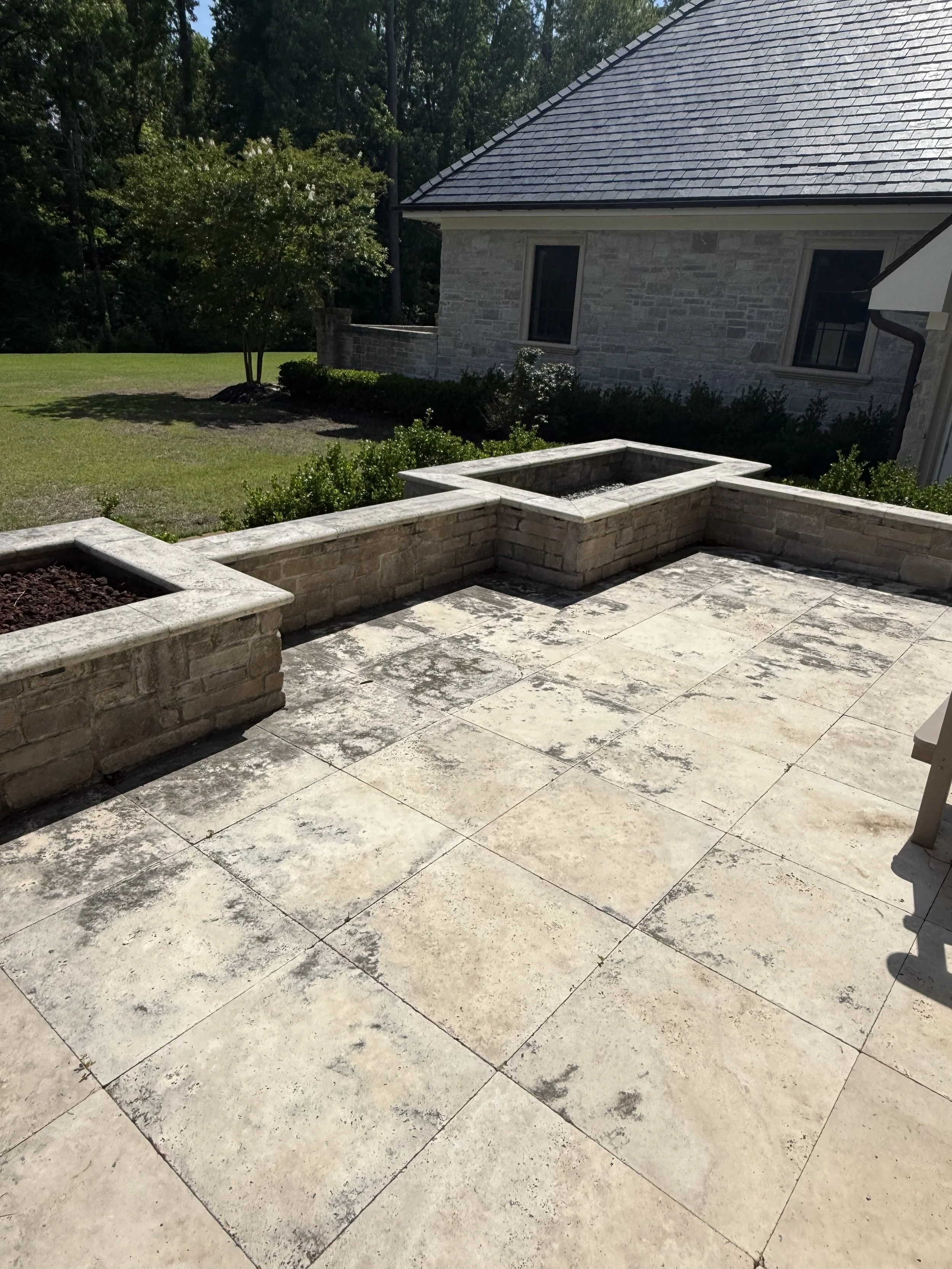 A backyard patio with large stone tiles, beige in color, and a stone wall with three raised planters, one with soil. There is a grassy lawn with a small tree and shrubs, with a house featuring a stone exterior, a sloped roof, and windows in the background. Dense trees and foliage are behind the house.