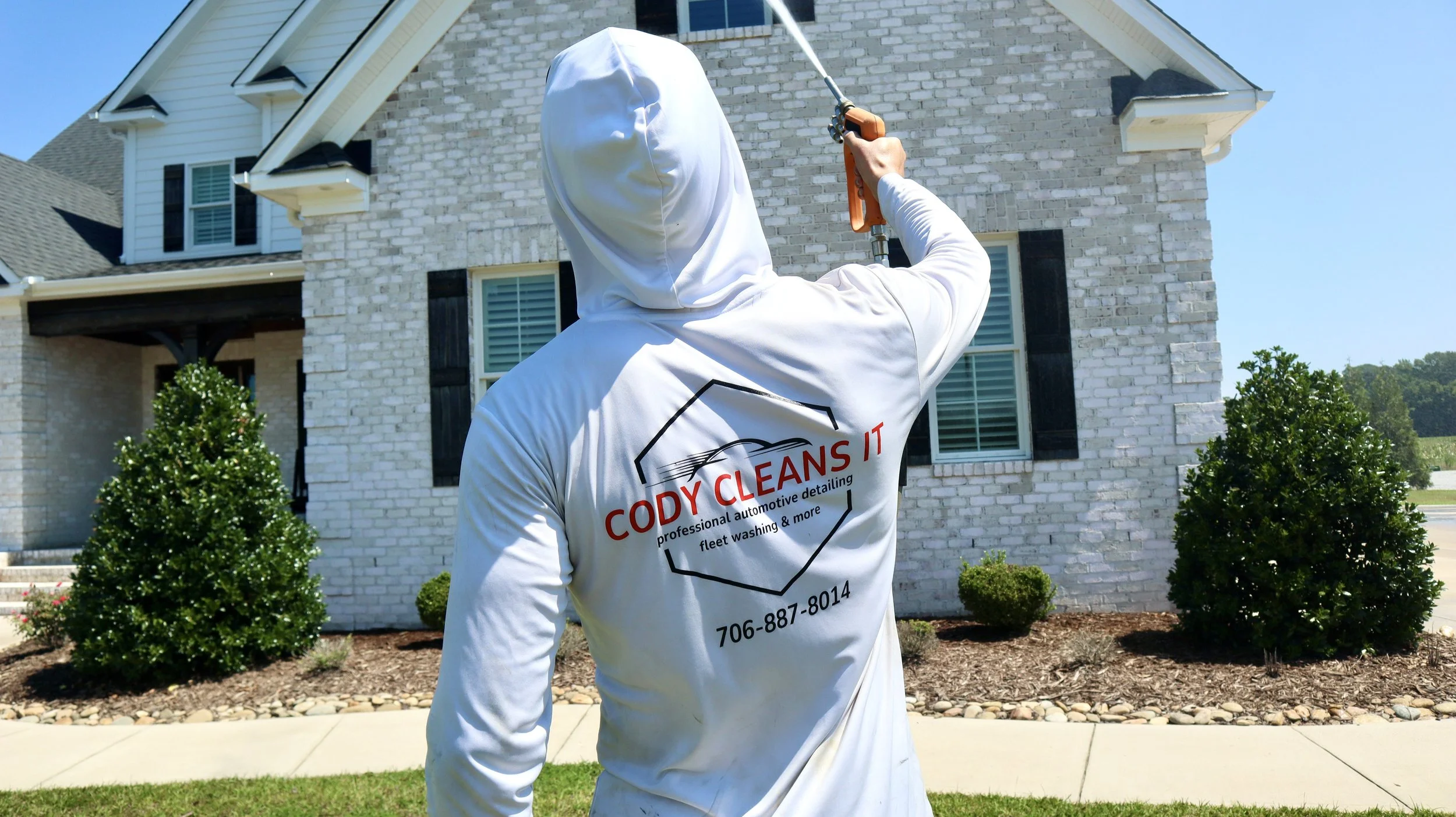 A person in white cleaning overalls facing a house, holding a power washer. The overalls have the business name 'Cody Clean It' and contact information on the back.