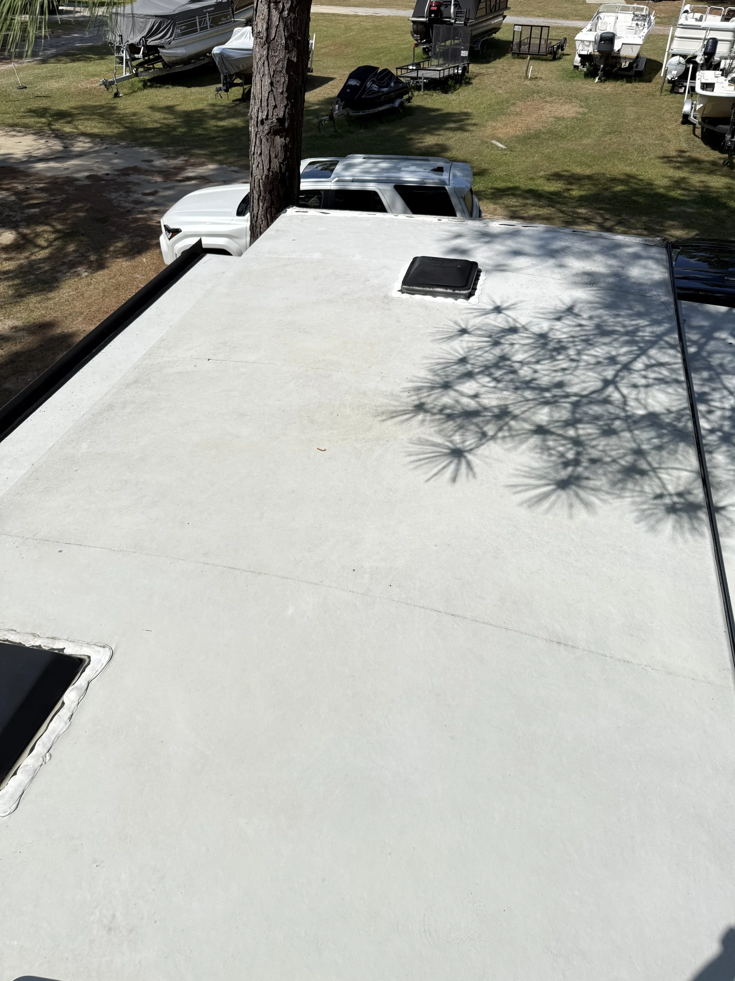 View from the top of a recreational vehicle looking down on a yard with parked boats, a large tree trunk, and outdoor equipment.