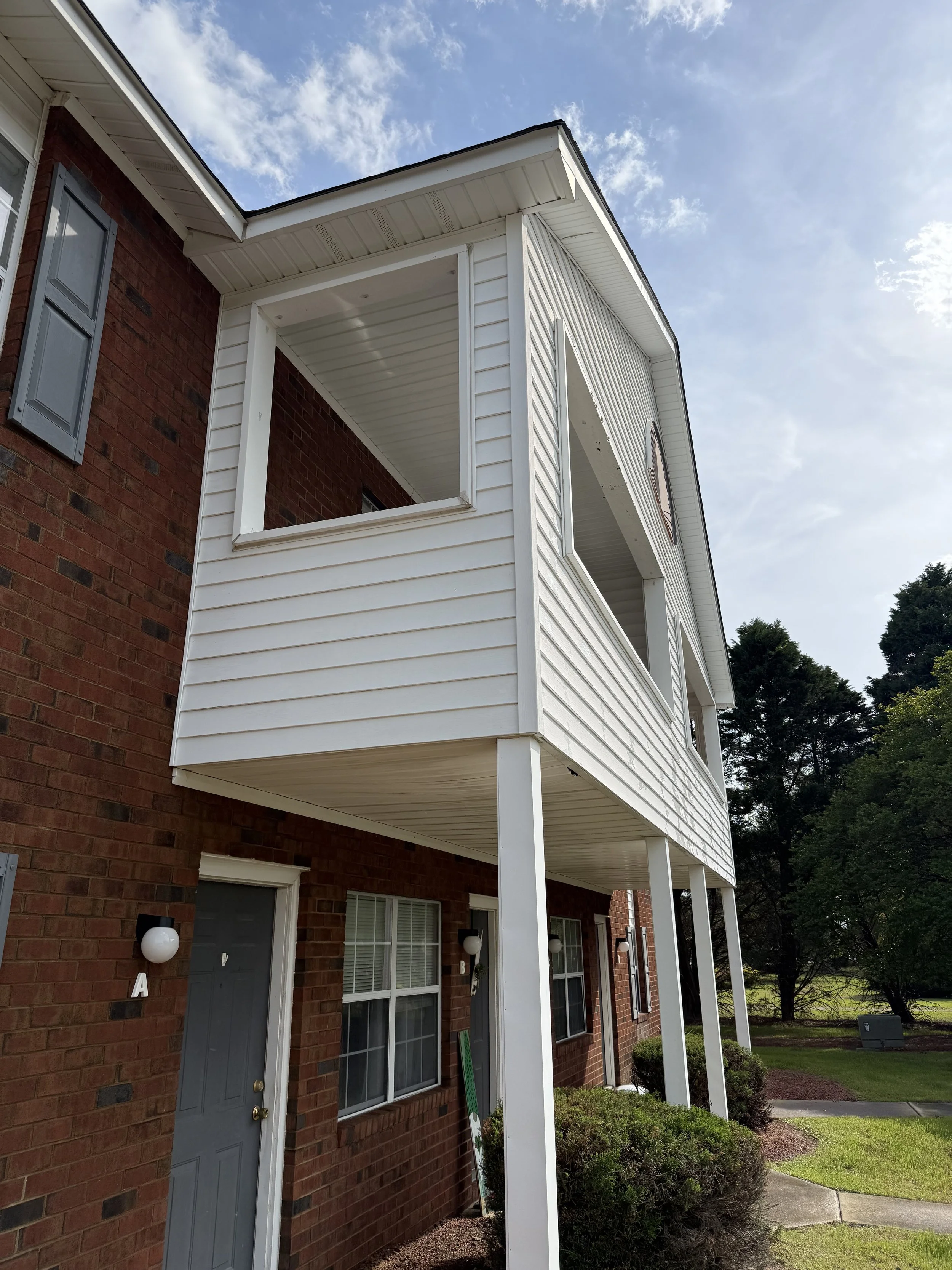 The image shows the exterior of a brick apartment building with a covered balcony and white siding. There are multiple windows with white blinds, spherical wall-mounted lights, and alphabetical labels on the doors. The scene includes a grassy yard with bushes and trees, under a partly cloudy blue sky.