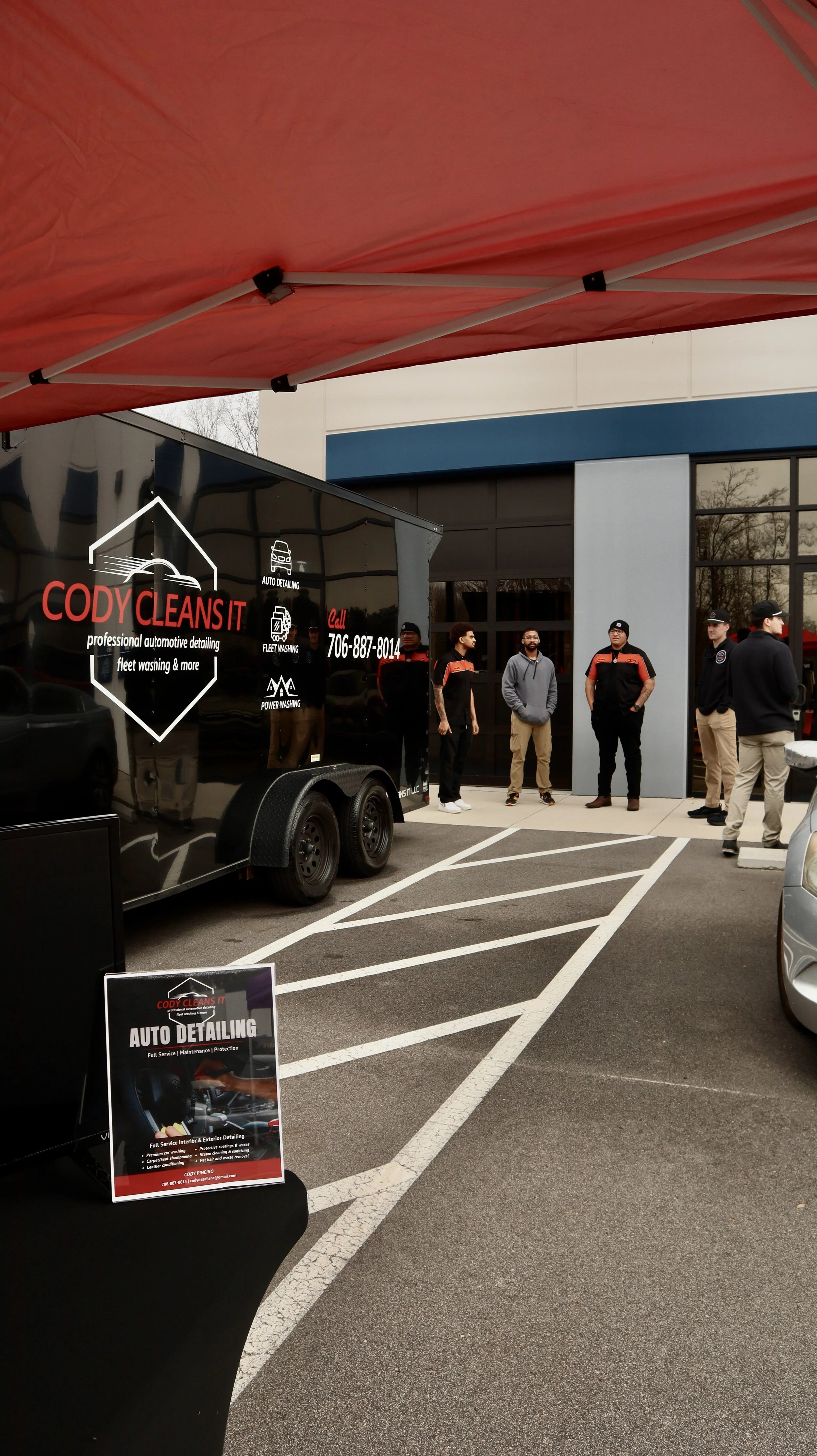 A group of people standing outside a building under a red canopy near a black mobile auto detailing trailer with signage that reads Cody Cleans It, specializing in auto cleaning services. There are parking lines visible in the foreground and a poster advertising auto detailing services.