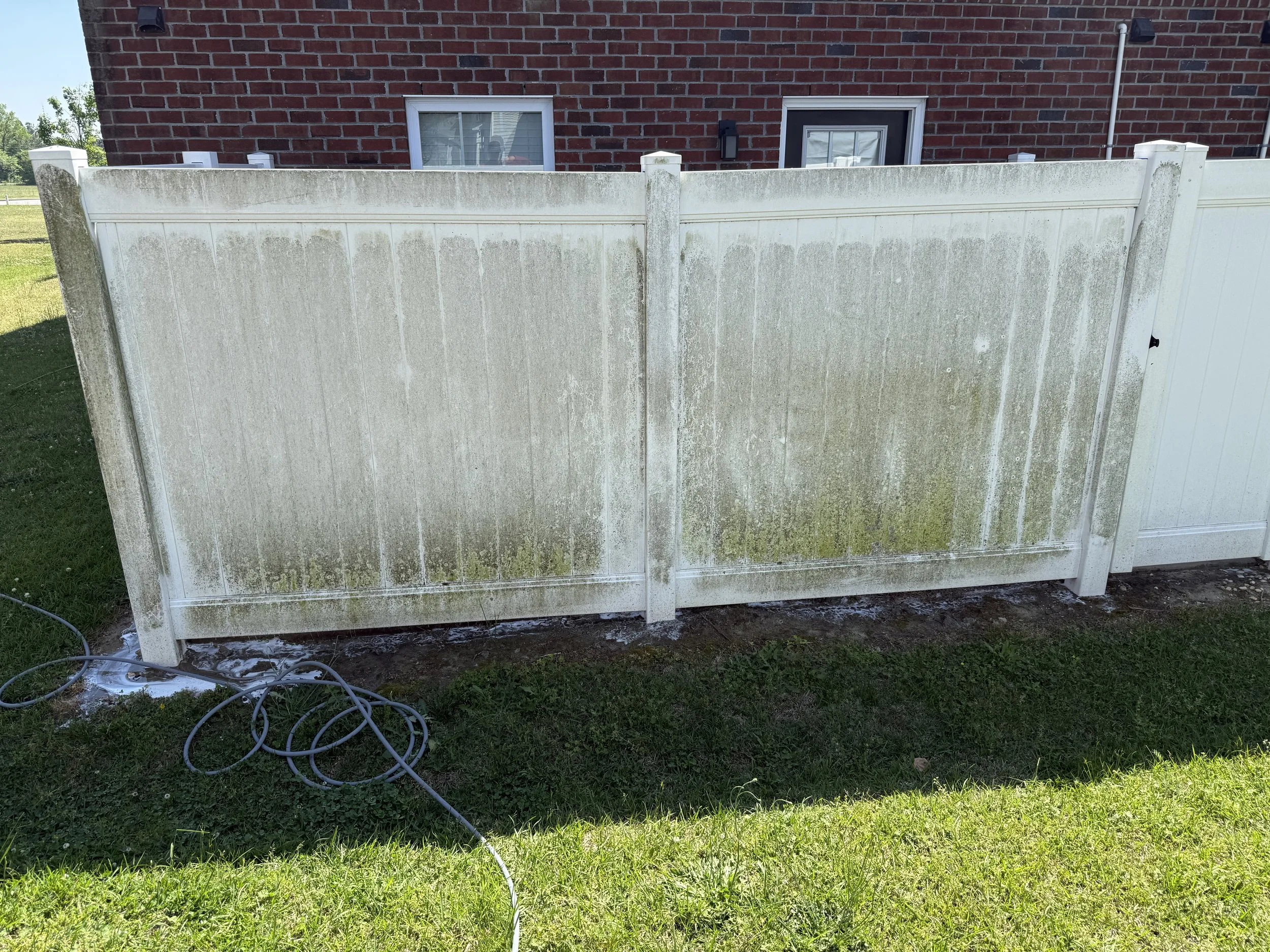 A white fence with moss and dirt stains in front of a brick house with two windows. A coiled garden hose is lying on the grass near the fence. The house has a brick facade and a black door with a small window.