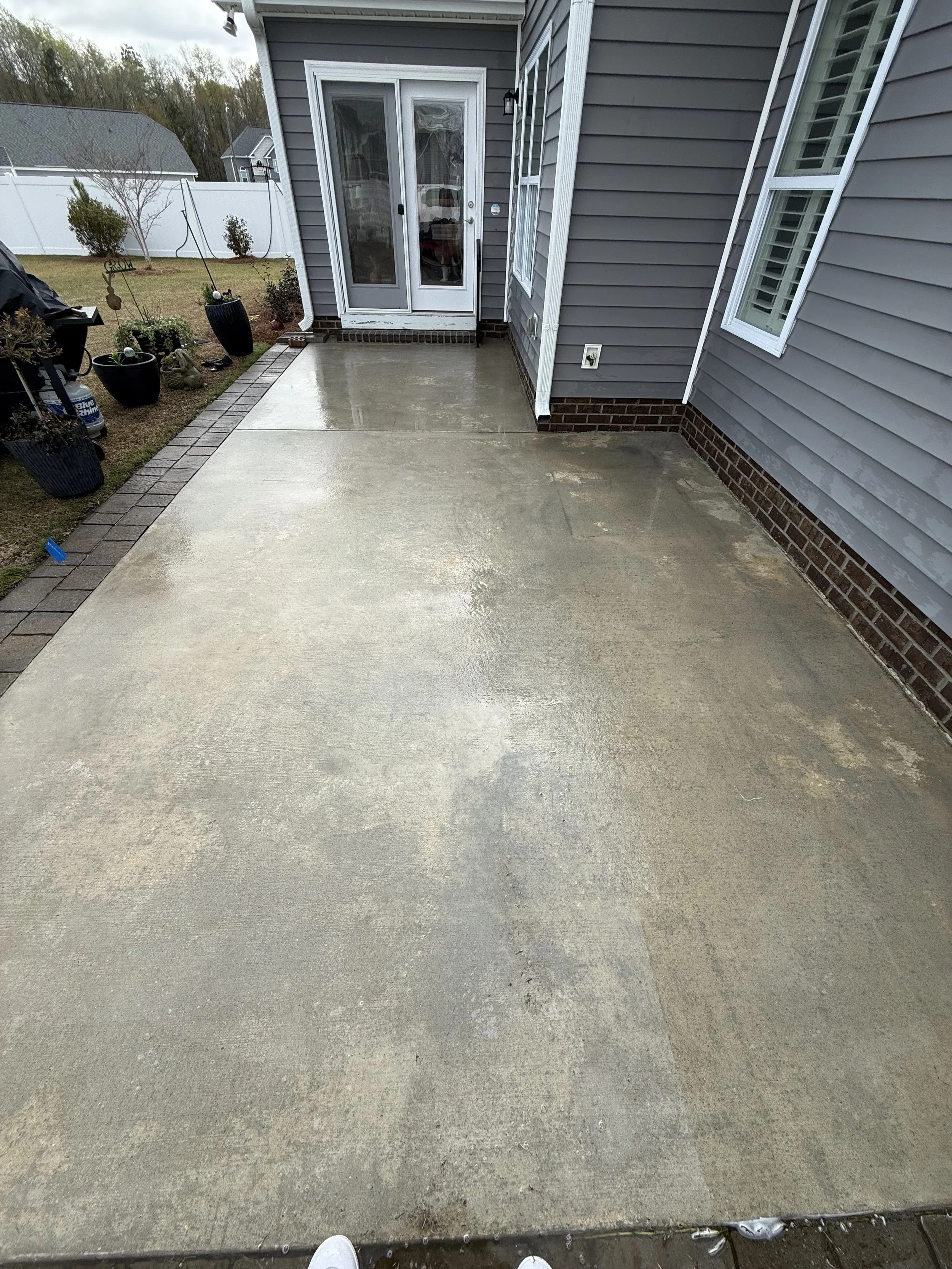 A wet concrete backyard patio in front of a house with gray siding and white trim, with some plants and decorations on the side.