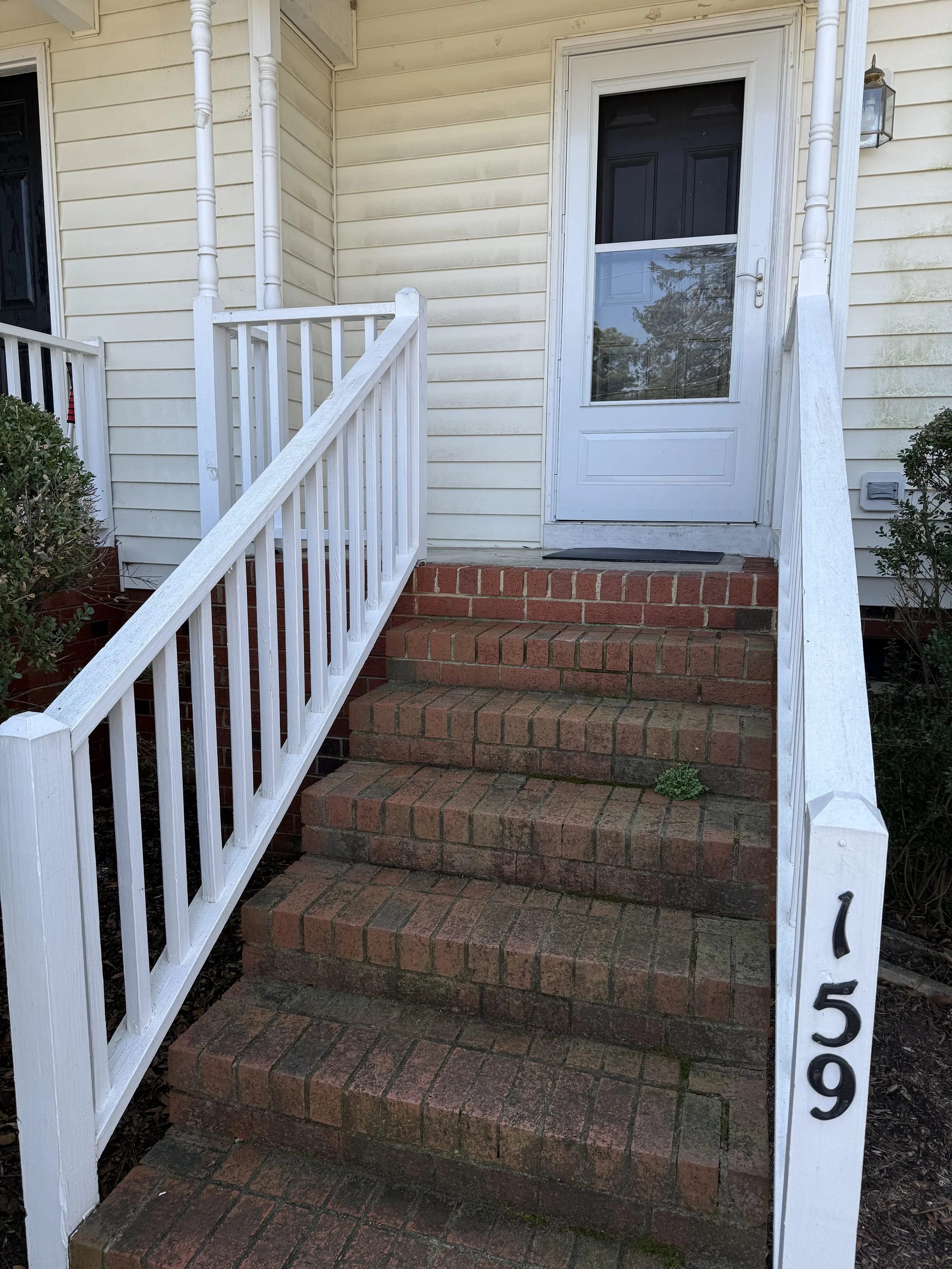 Front porch of a house with brick stairs, white railing, and a white door with a screen. House siding is light yellow with some dirt or discoloration. The house number '509' is displayed vertically on the white post.