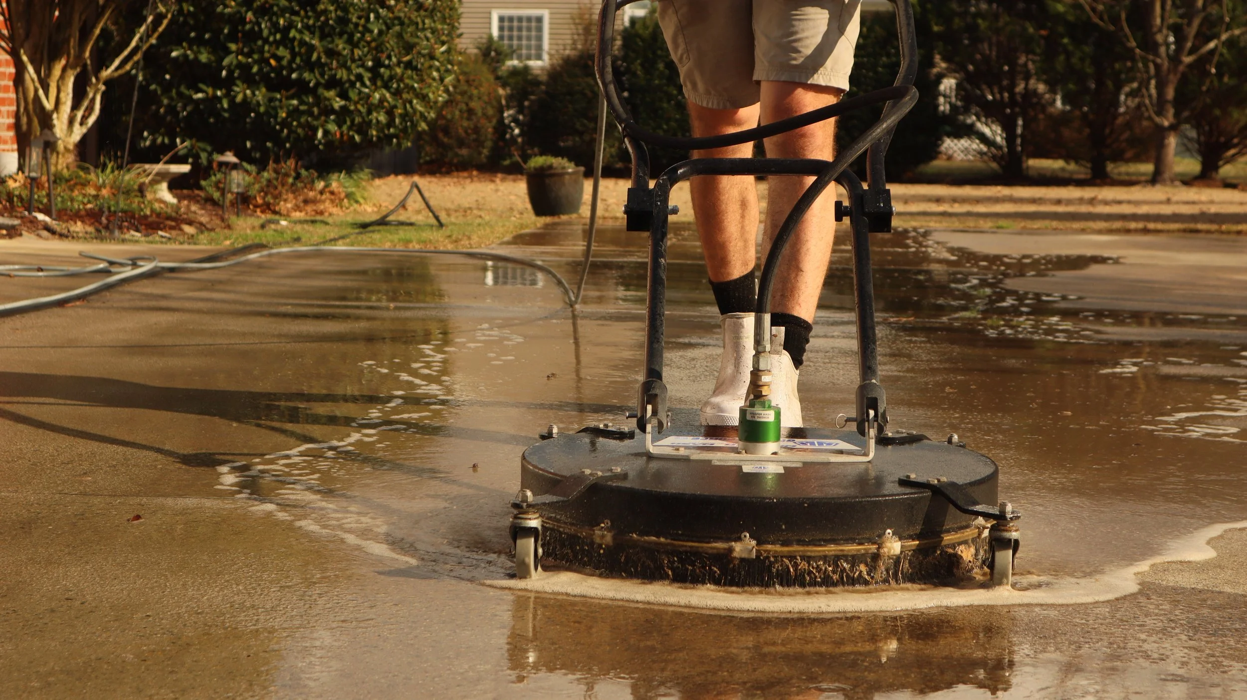 Person in shorts and boots pressure washing a concrete driveway with a large power washer, water streaming across the surface.