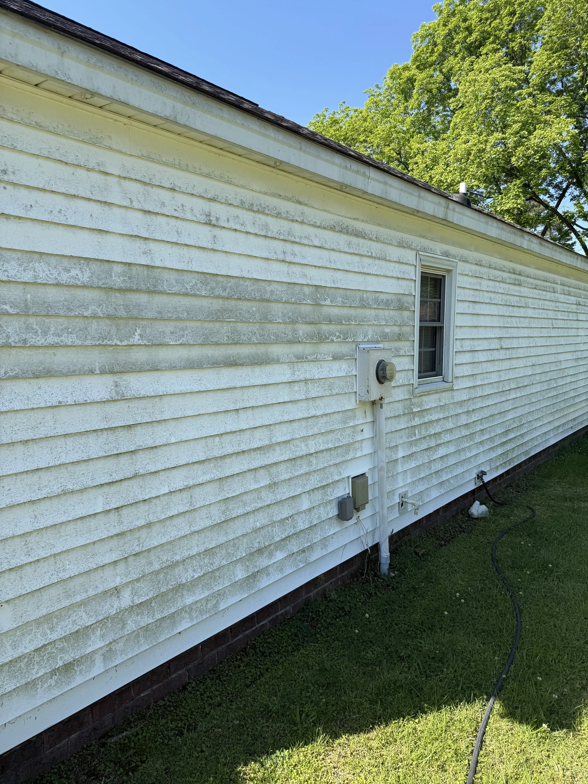 The side of a house with yellow siding showing signs of weathering and dirt, a small window, and outdoor electrical meters and wires underneath the window. There is a grassy yard and a large green tree with a blue sky in the background.