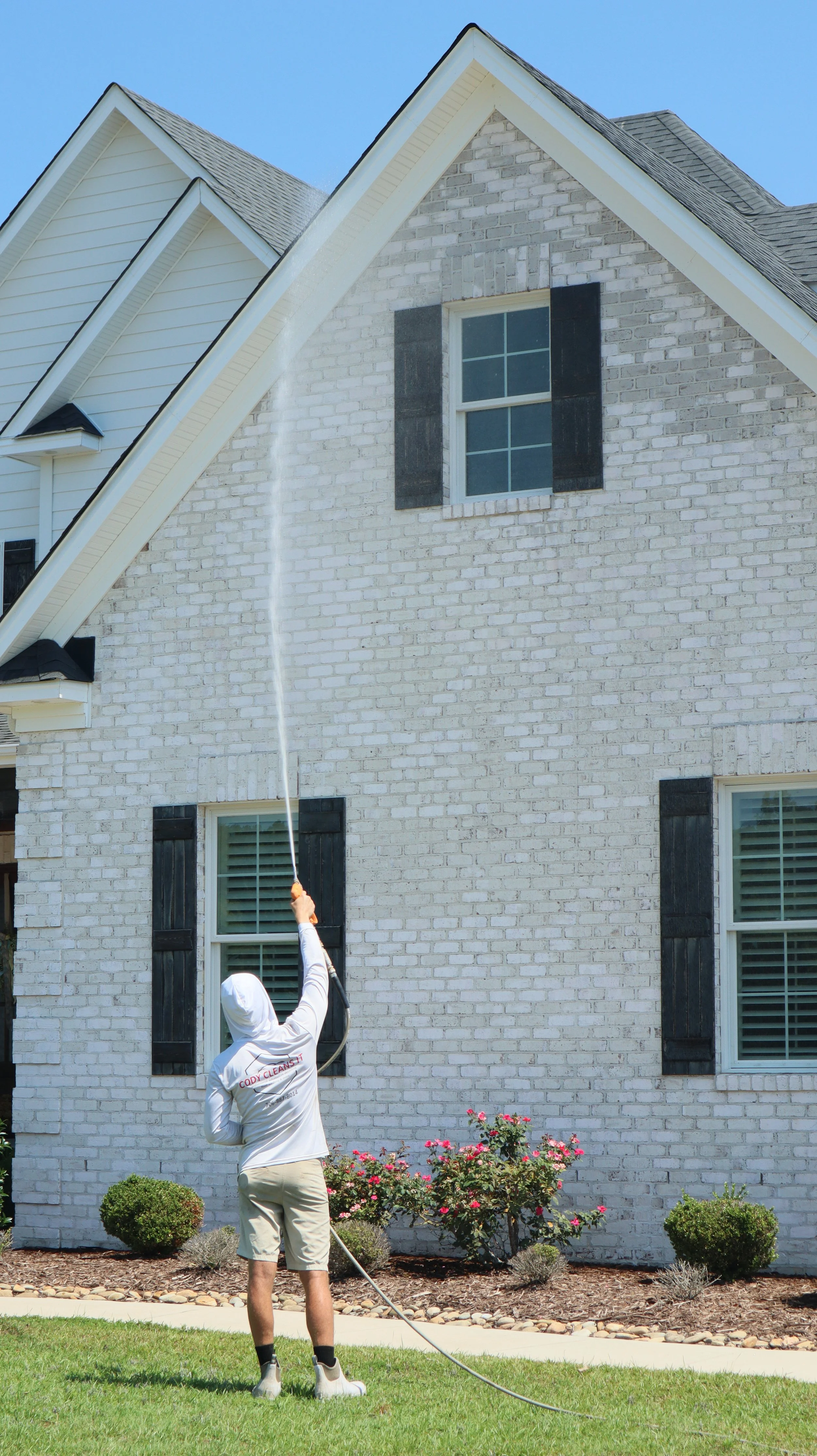 Person wearing a hoodie and shorts pressure washing the side of a white brick house with black shutters.