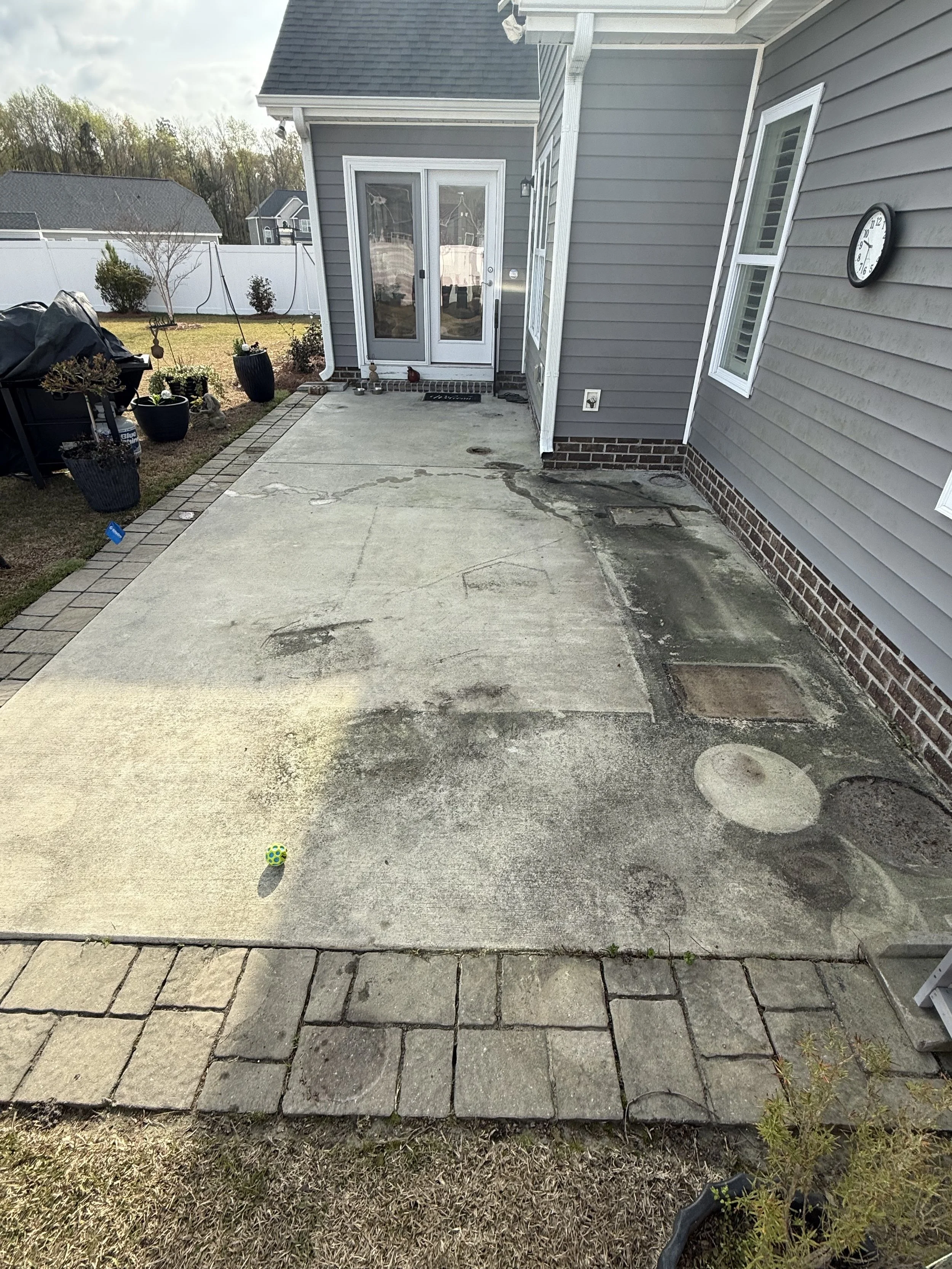 Backyard with concrete patio, potted plants, hanging planter, glass sliding door, windows, wall clock, and a small ball on the ground.