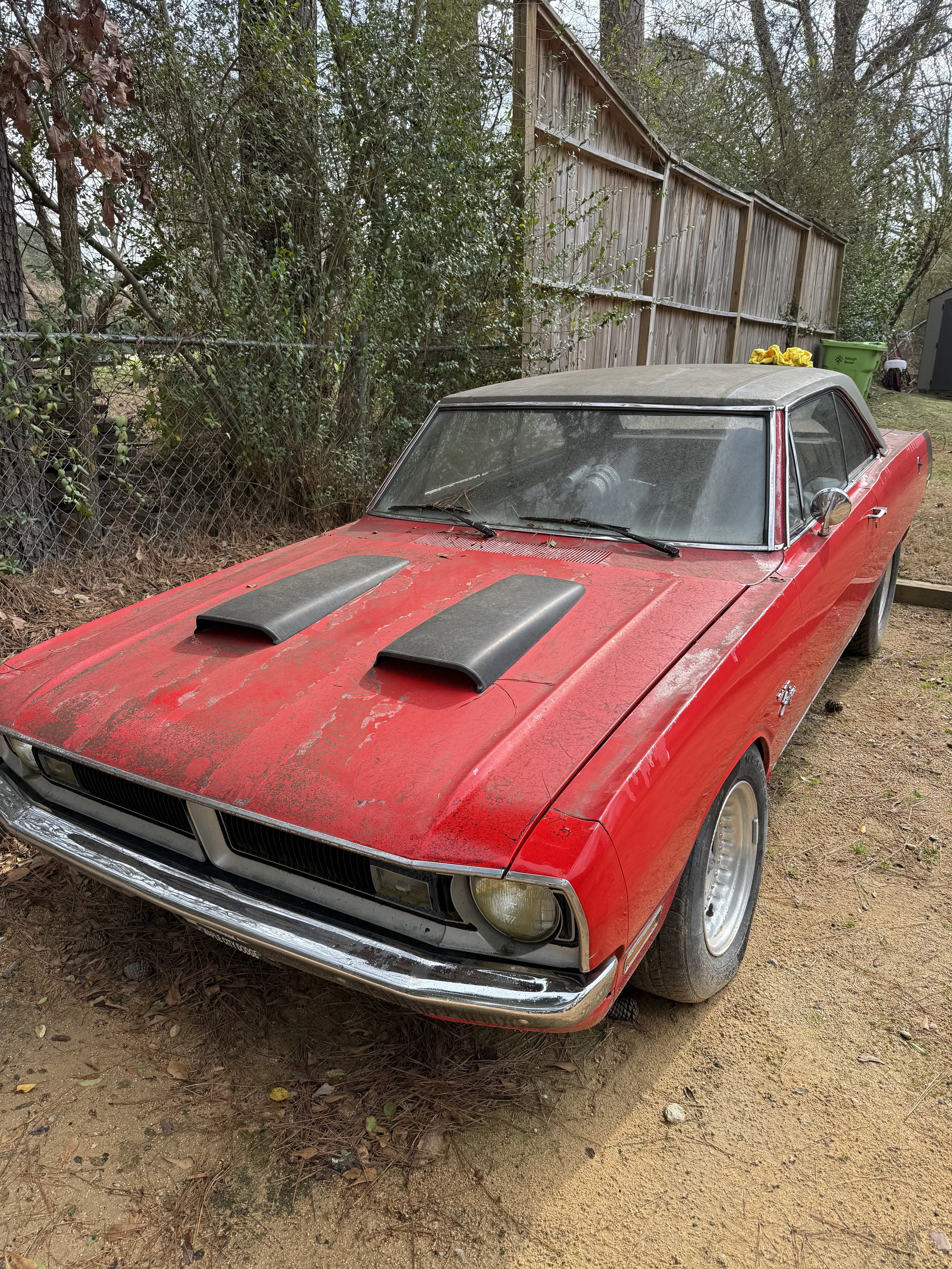 A red vintage sports car with black hood vents parked outdoors on dirt ground, dusty and dirty windshield, old-aged look, surrounded by trees, a wooden fence, and some trash cans in the background.